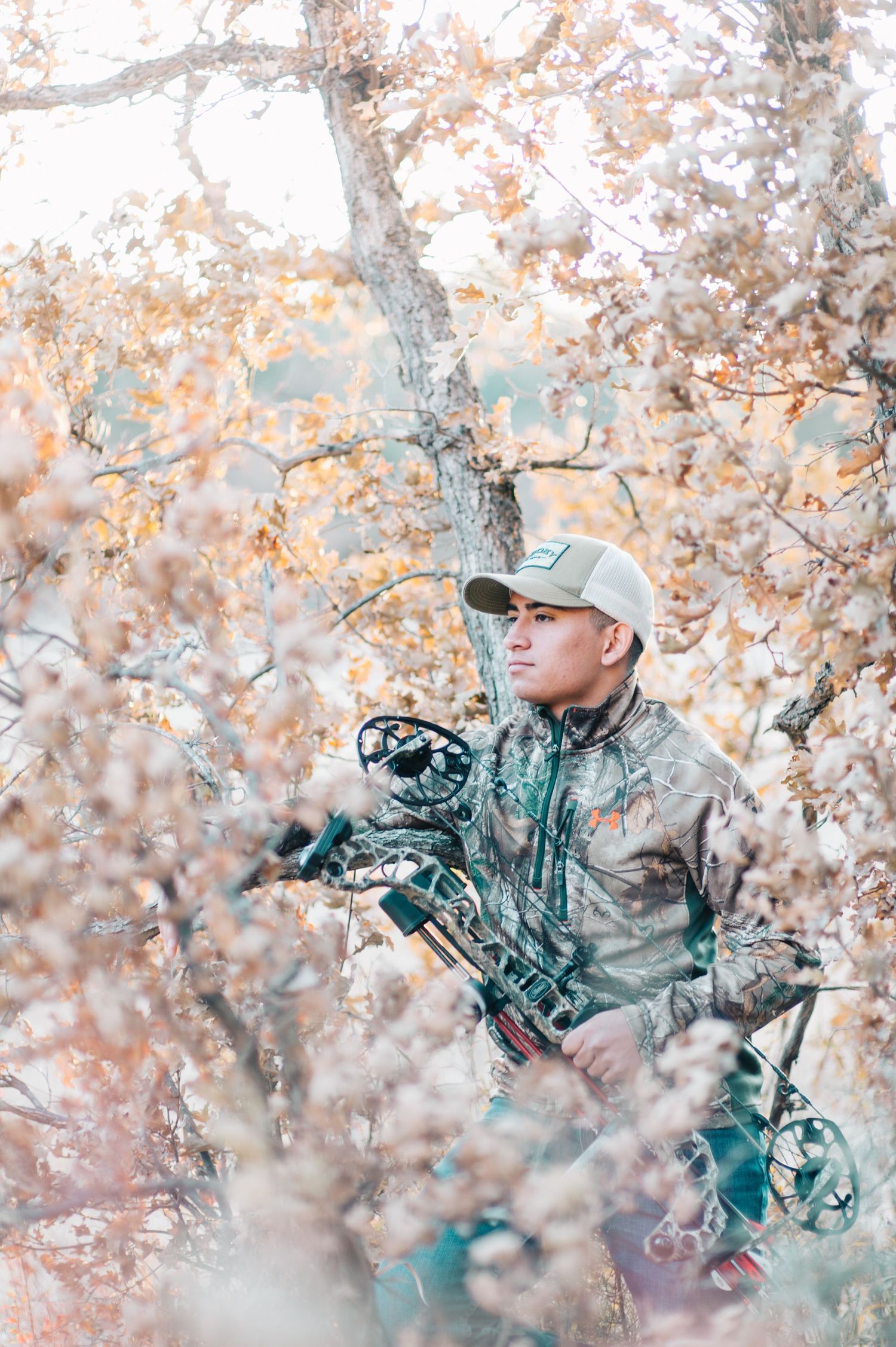 Hunter in camo gear holding a compound bow while standing among autumn trees.