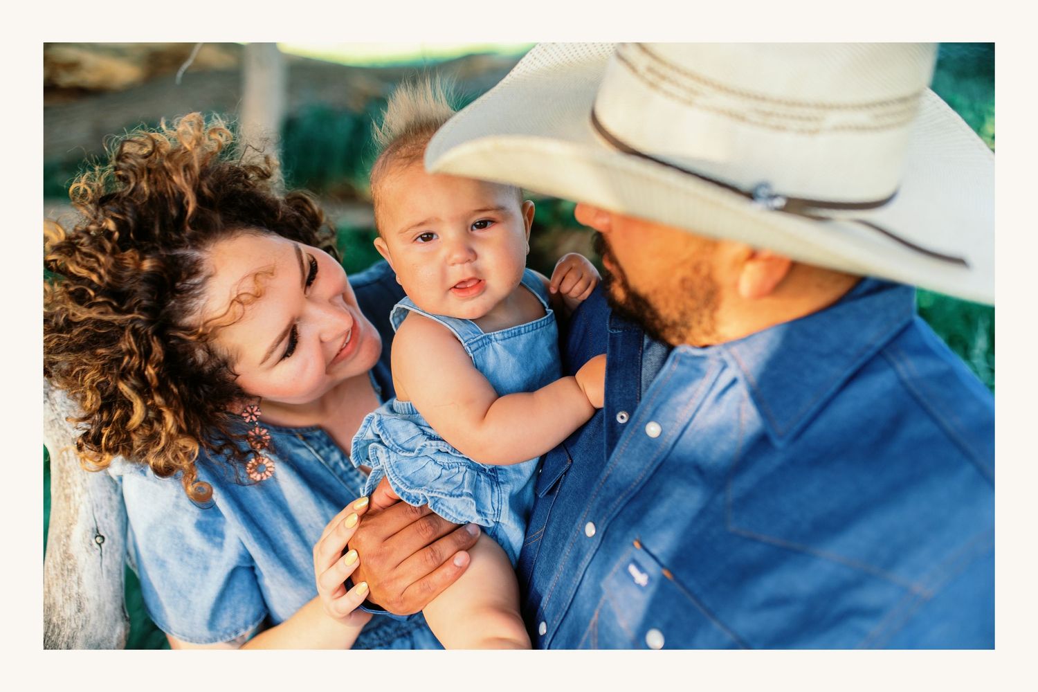 A young family in denim shirts, mother holding baby girl, father in cowboy hat, smiling outdoors in warm sunlight.