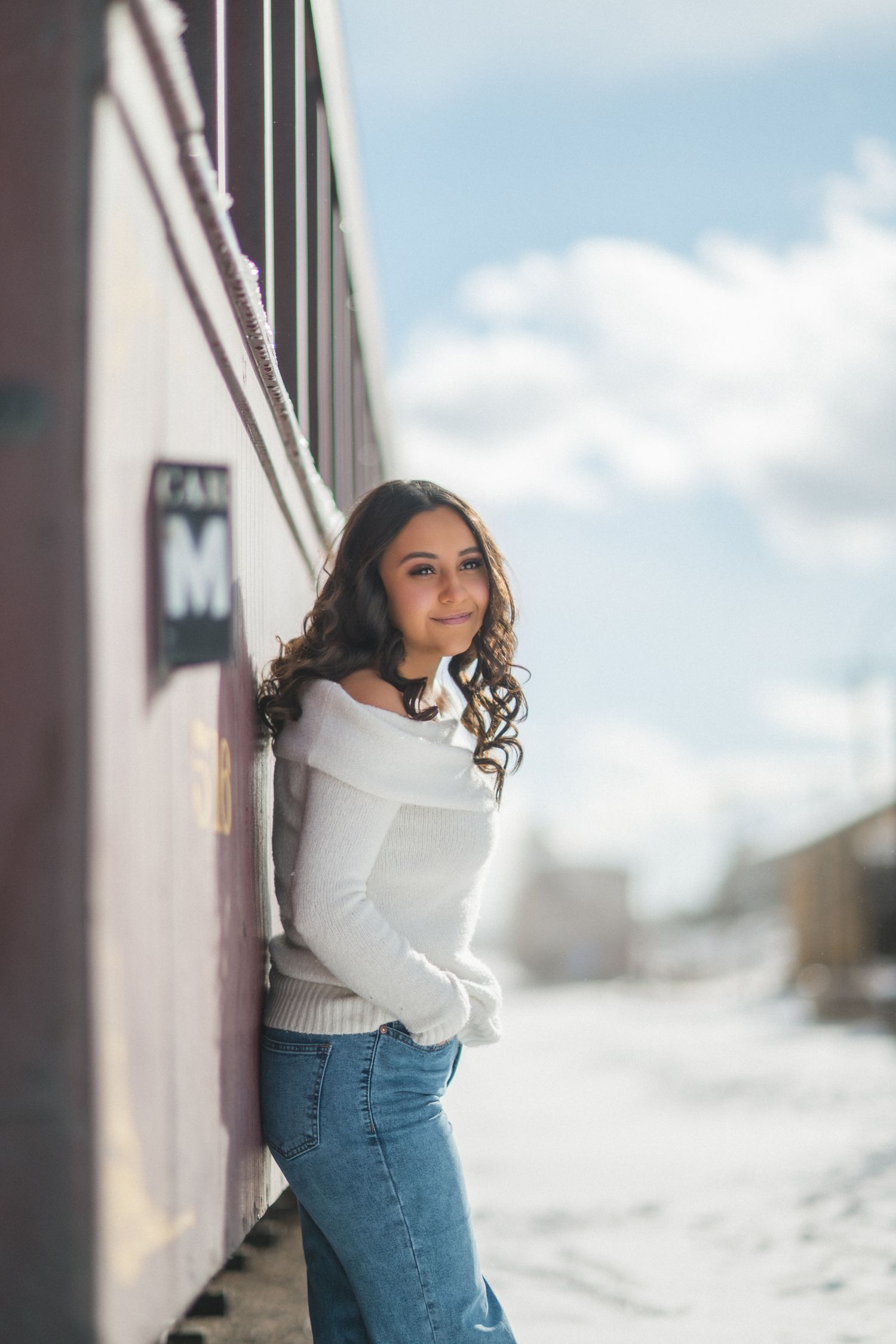A person in a white sweater and jeans poses against a wall outdoors during winter.
