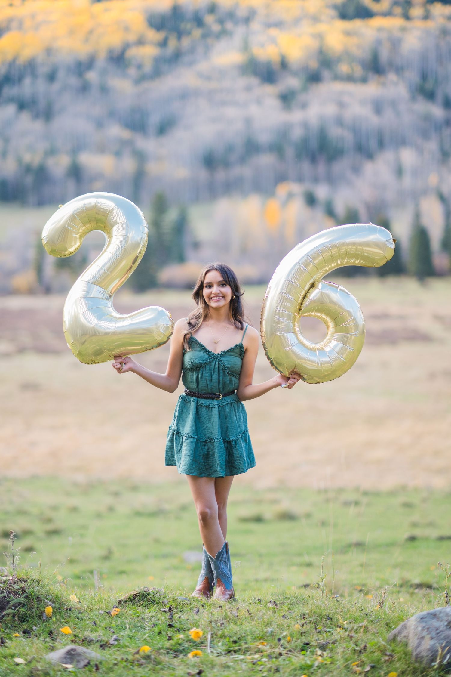 A person in a teal dress holds large metallic letter balloons in an autumn field with mountains in the background.