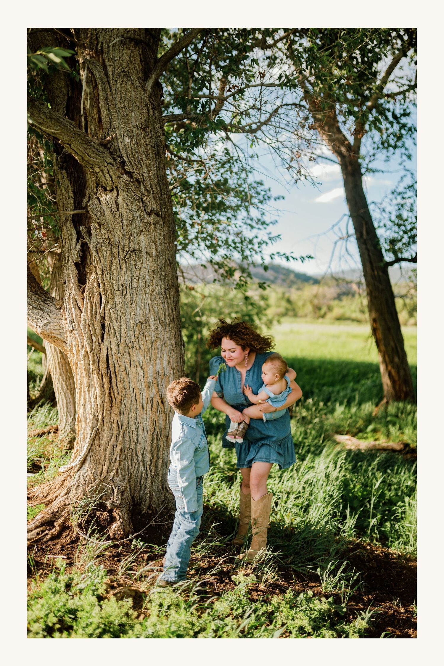 A mother holds her baby while interacting with her young son beside a large tree in a lush green outdoor setting.