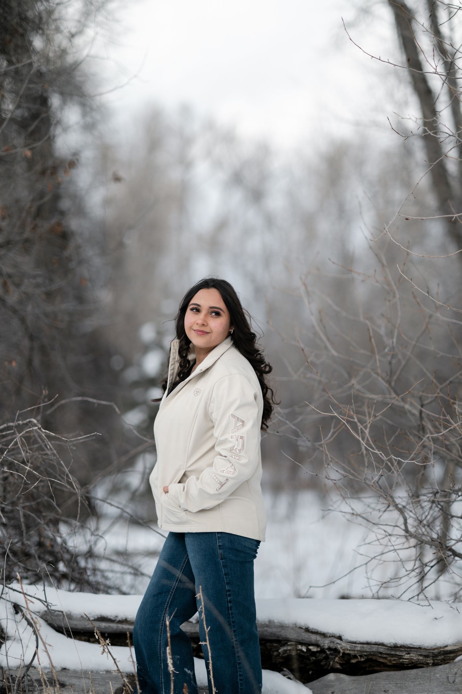 A winter portrait session in a snowy forest setting with someone wearing a white sweater and jeans.