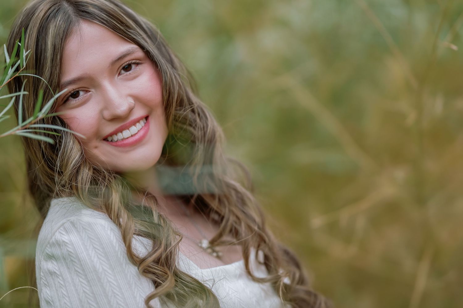 A person with brown wavy hair sits in a grassy field wearing casual clothes and a warm smile.