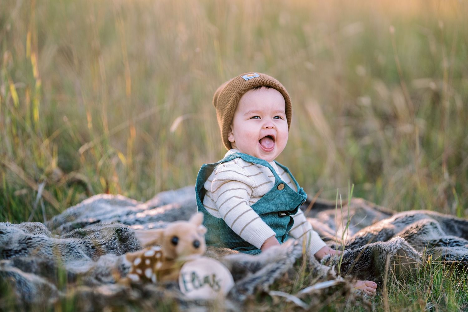 A baby in a brown hat sits on a blanket outdoors, surrounded by natural scenery.