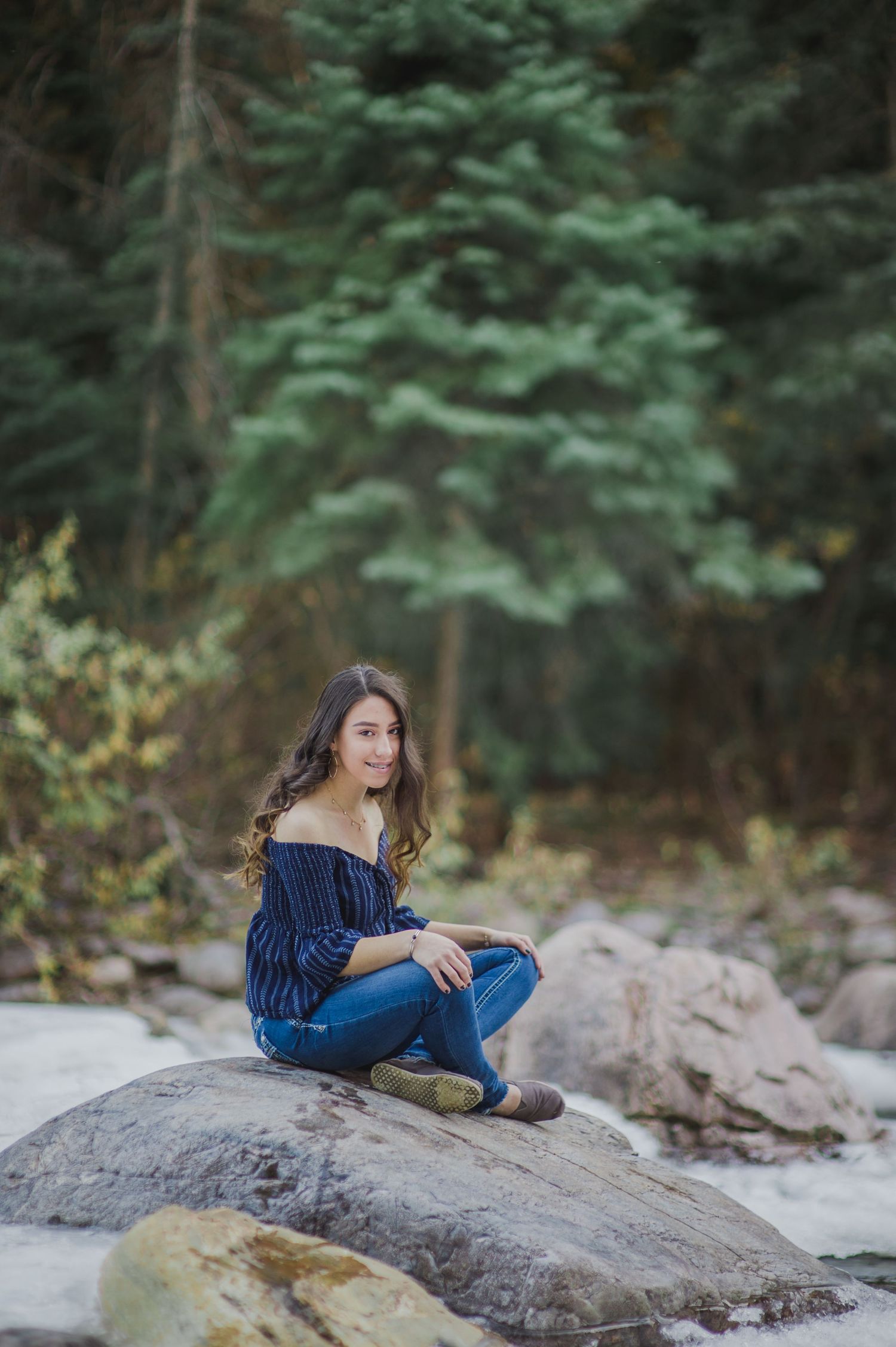 Young woman in blue off-shoulder top and jeans sitting on rocks near a stream surrounded by evergreen trees.