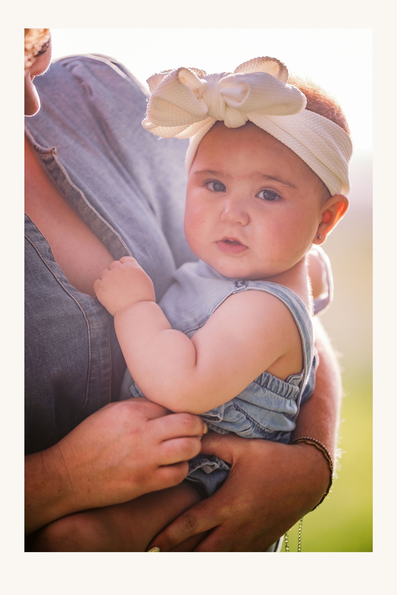 A baby girl in denim overalls and a white bow headband being held by an adult outdoors in soft sunlight.