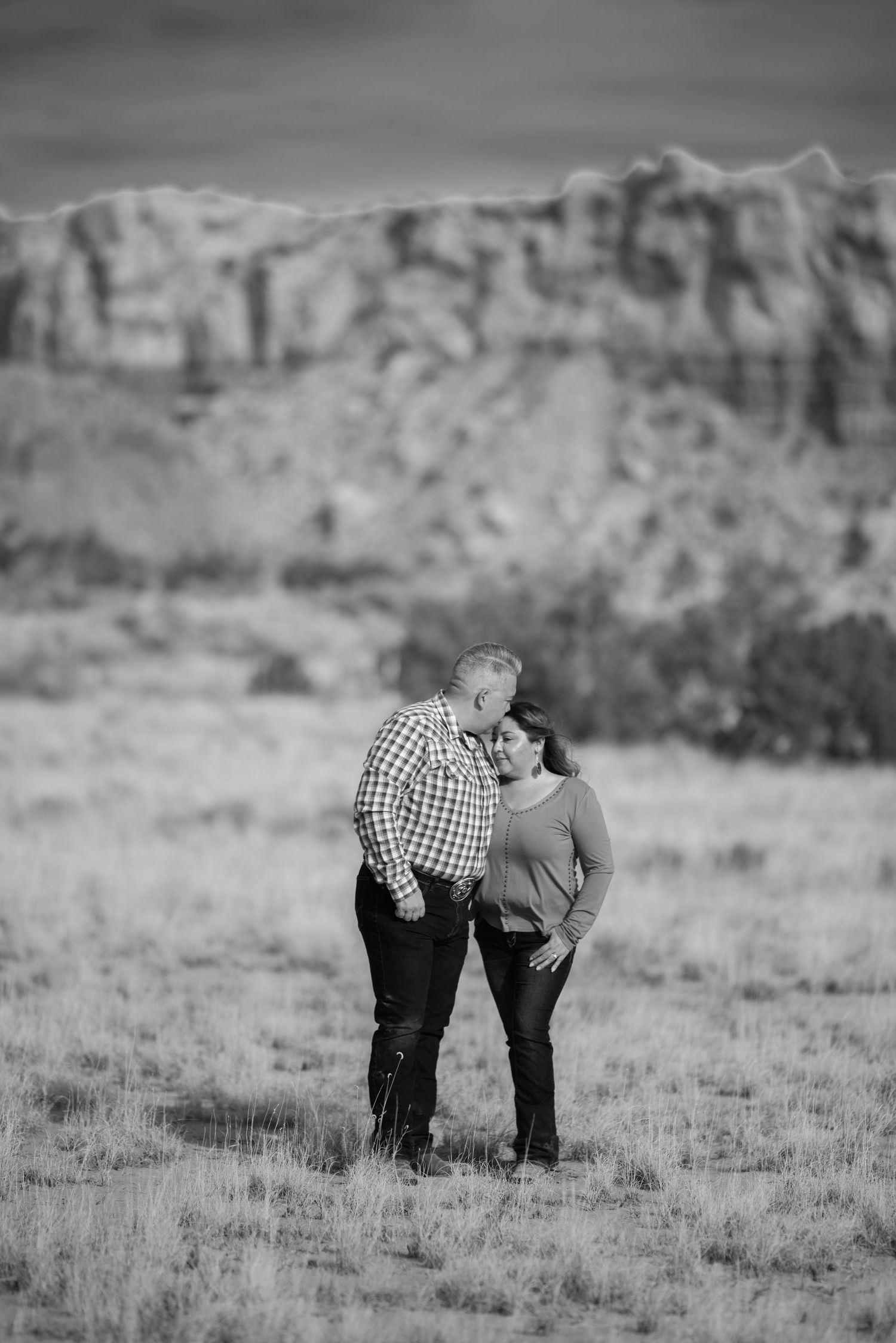 A couple shares an intimate moment in a field with dramatic desert cliffs in the black and white background.