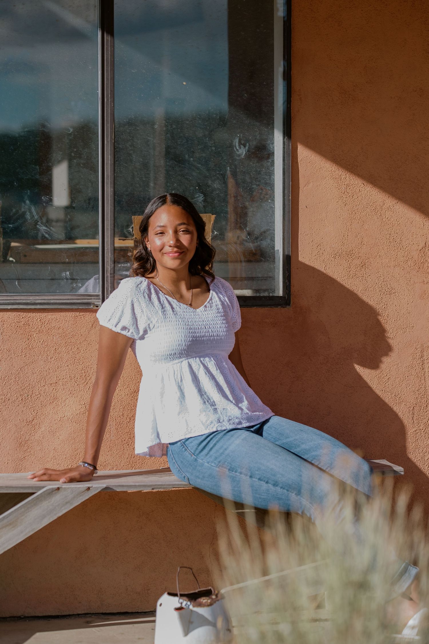 A woman in light clothing lounges and smiles on a porch bench against a terracotta wall with windows in sunlight.