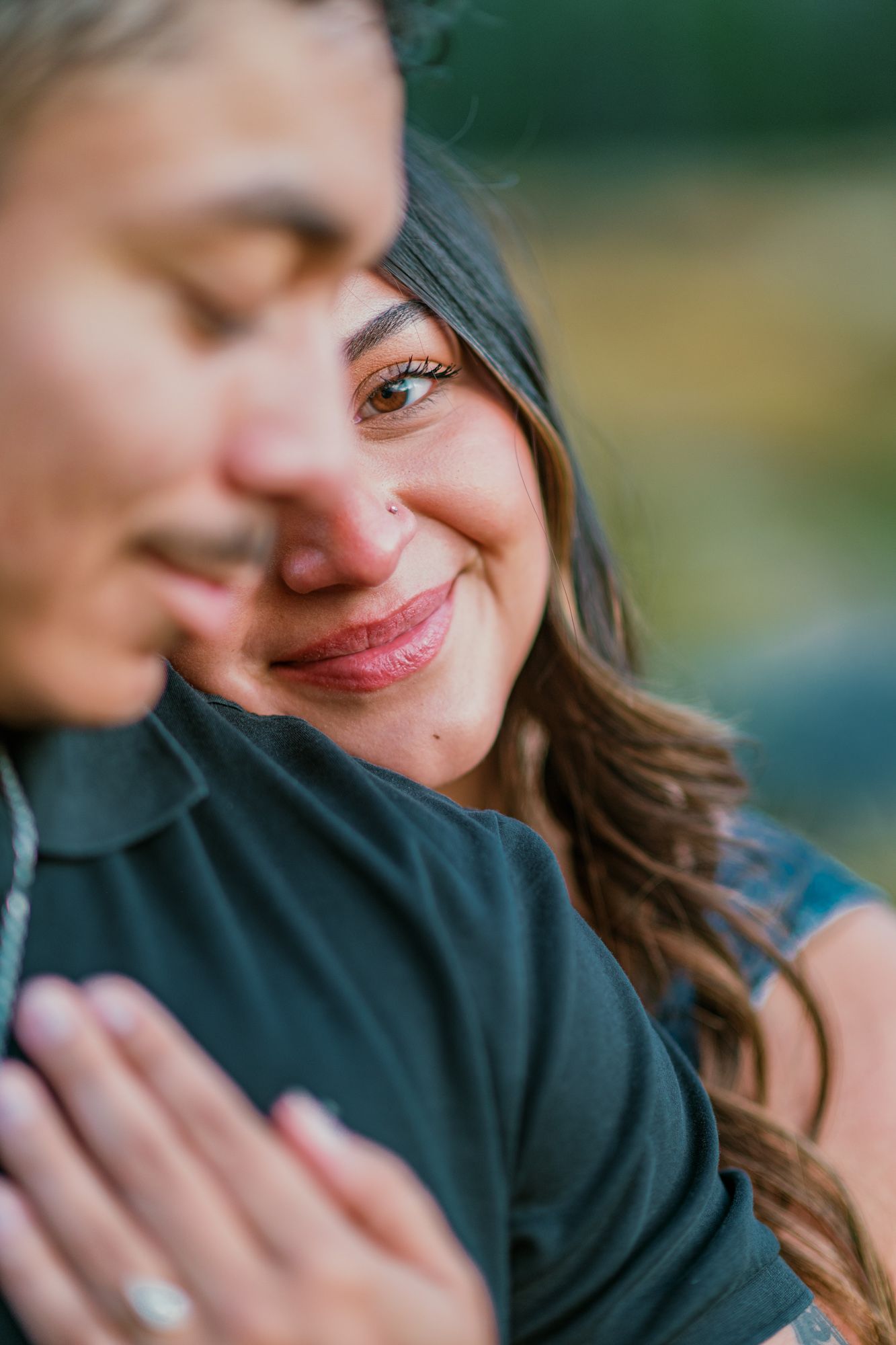 A woman smiles warmly while embracing a man from behind, her hands clasped on his chest in a romantic couples portrait.