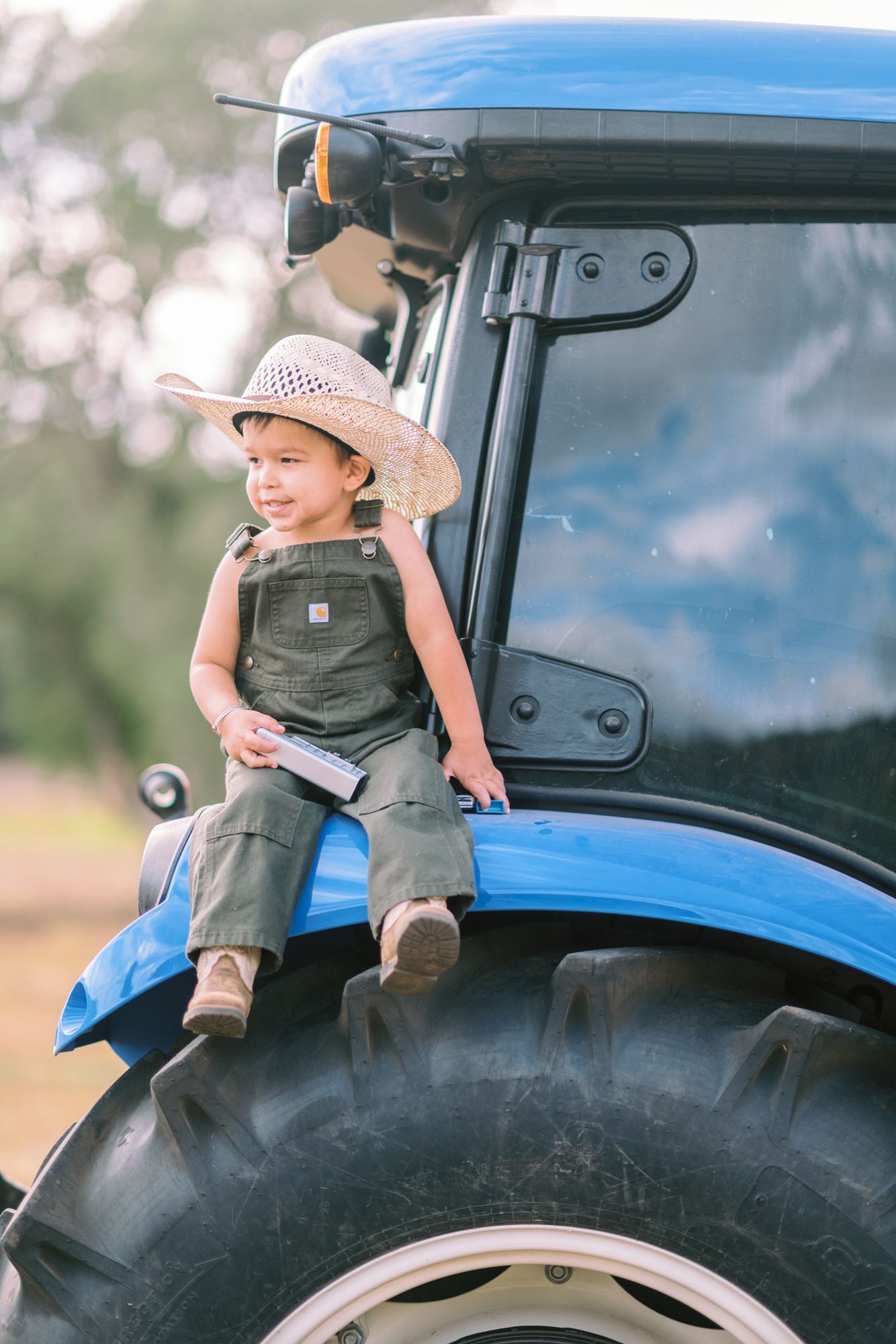 Young child in overalls and straw hat sitting on a blue tractor tire on a farm.