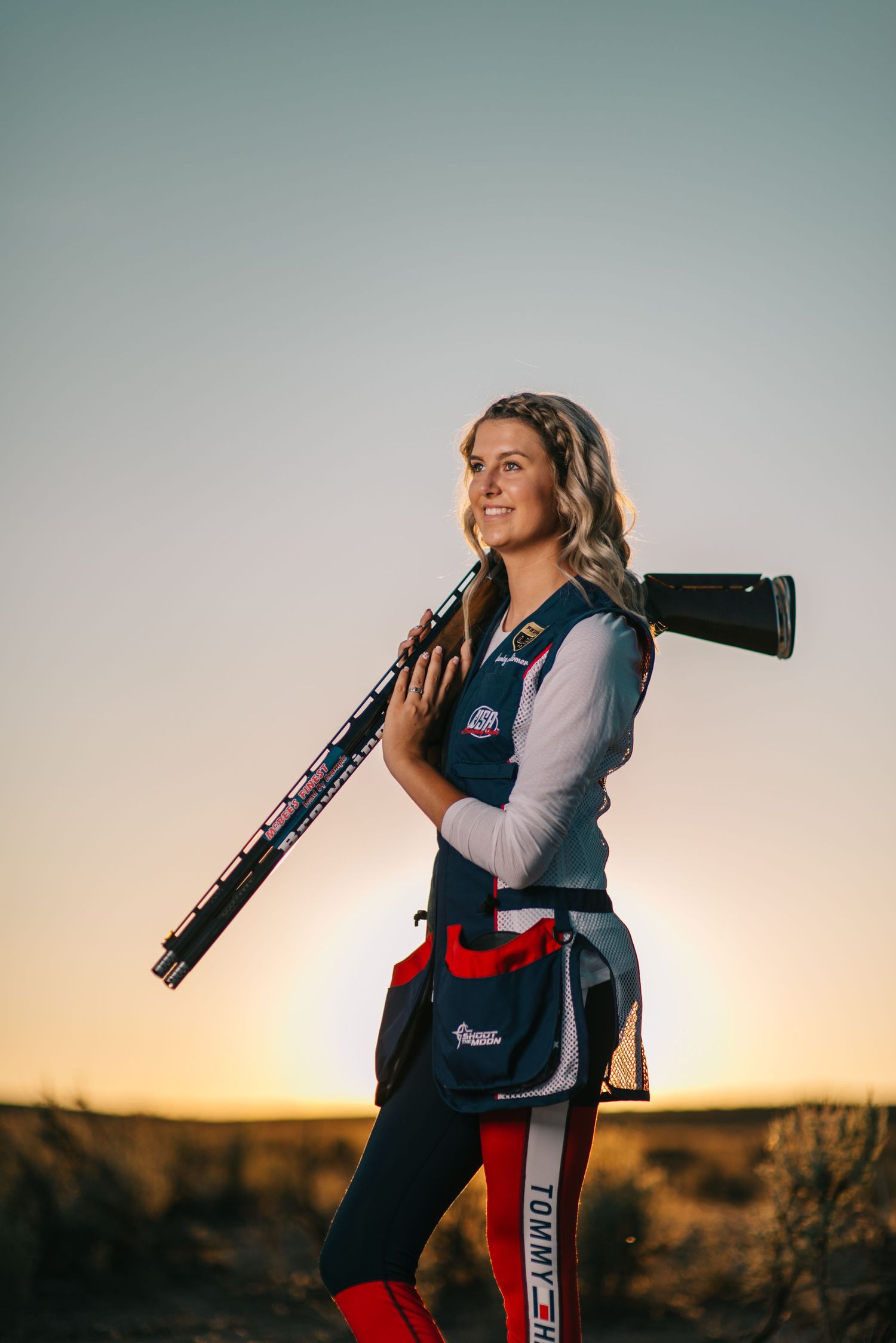 Competition shooter poses with rifle against sunset in professional shooting vest.