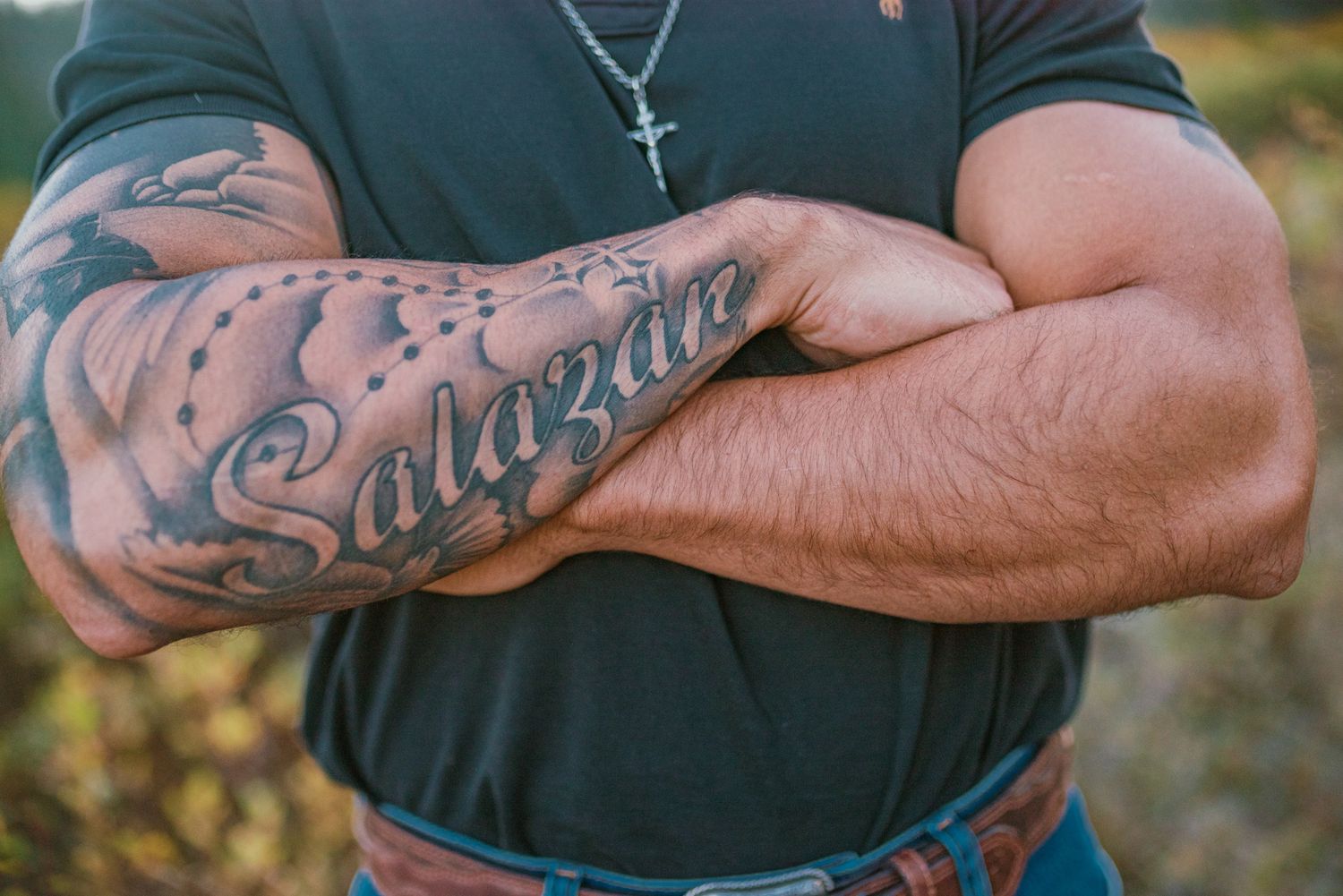 Muscular man with crossed arms showing a Salazar tattoo on forearm, wearing a cross necklace and dark shirt.