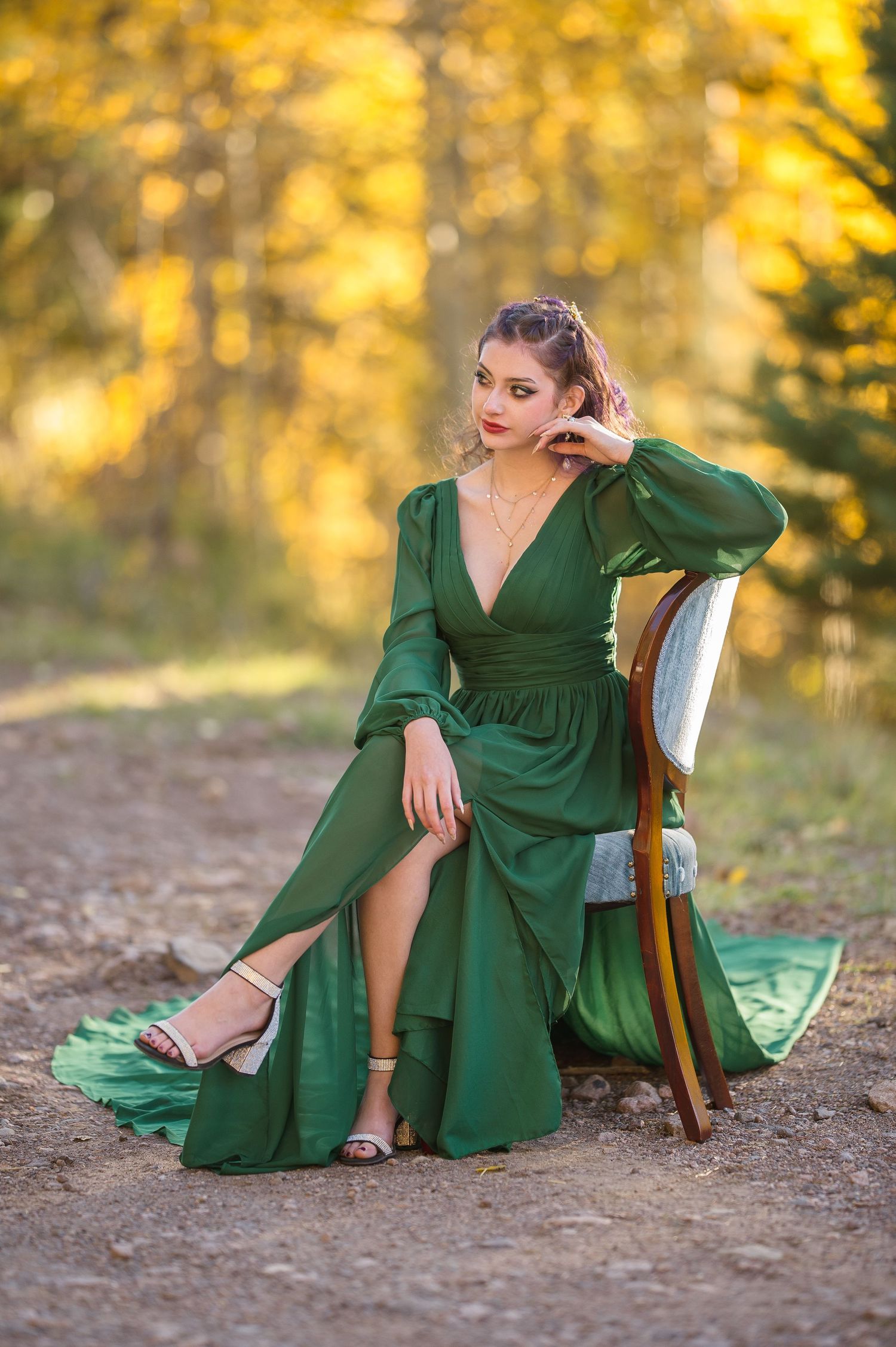 A person in an emerald green wrap dress poses elegantly on a wooden chair against a golden autumn backdrop.