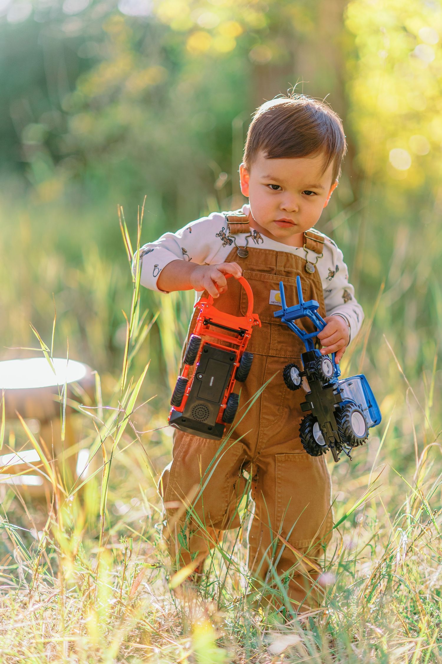 A small child wearing brown overalls plays with colorful toy trucks while sitting in tall grass on a sunny day.