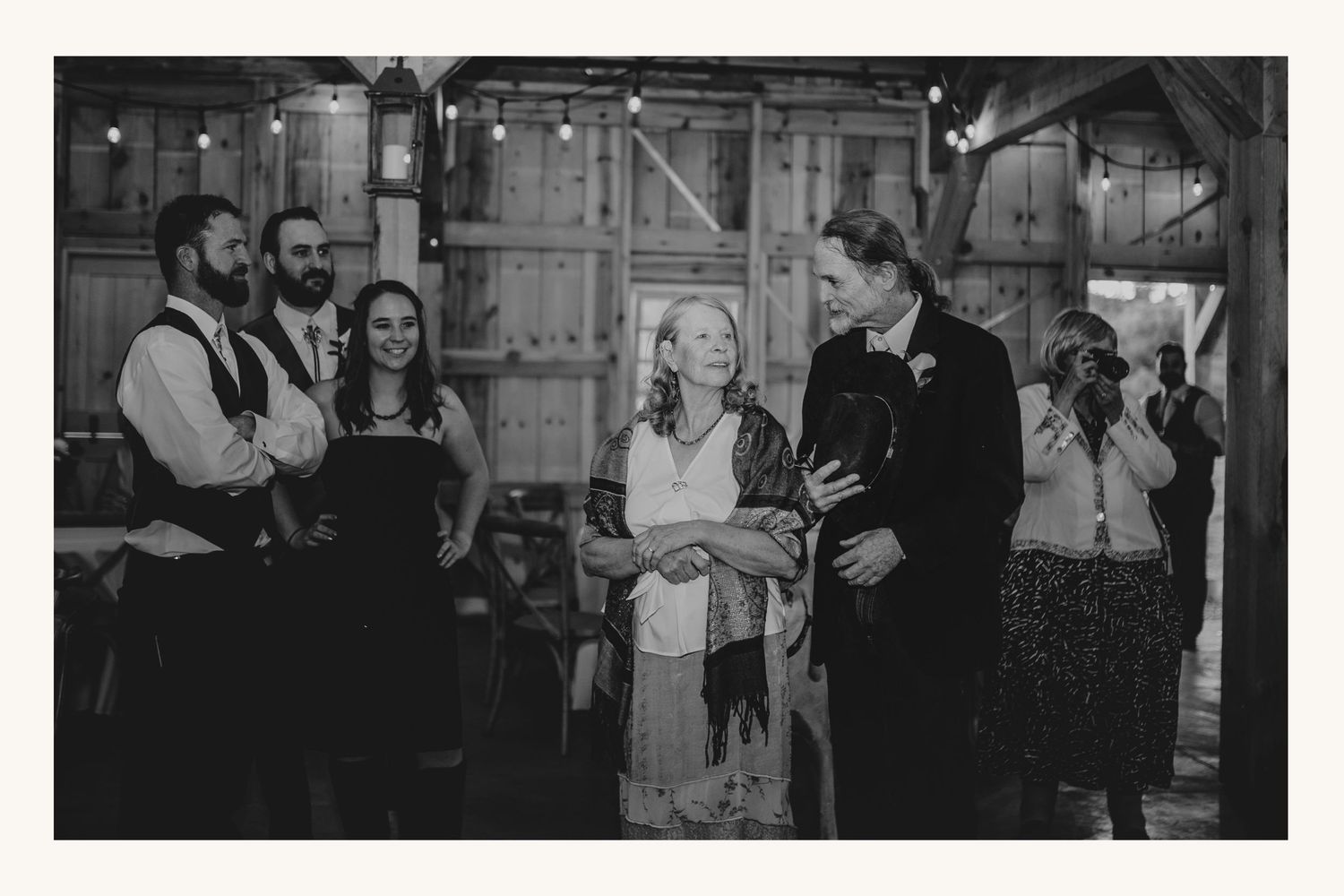 Black and white wedding reception photo of guests laughing and celebrating inside a rustic barn venue.
