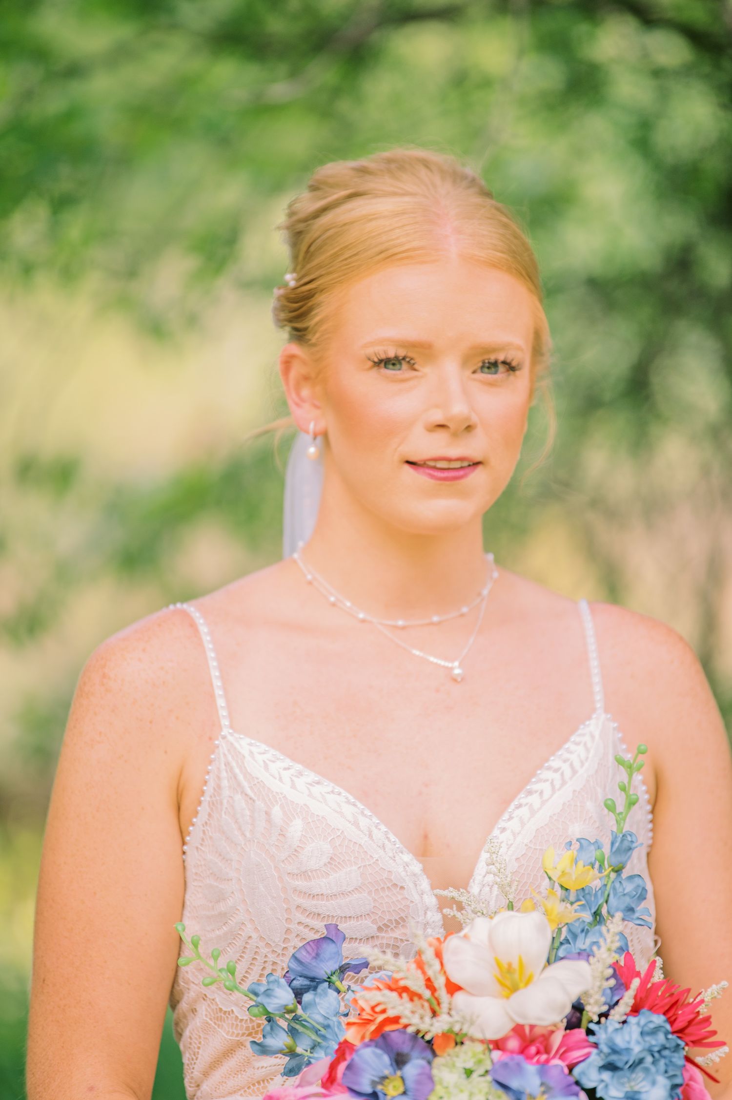 Bride in beaded gown holding colorful bouquet of blue and pink flowers, with pearl necklace and updo hairstyle.