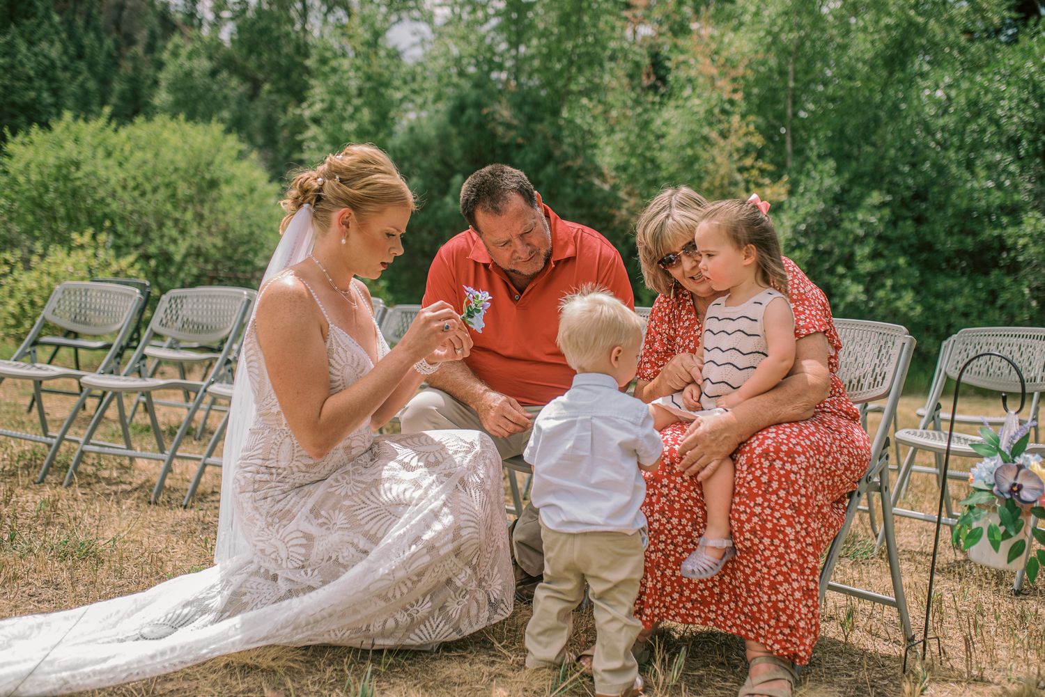Bride and groom sitting outdoors with two young children at a wedding ceremony surrounded by chairs and greenery.