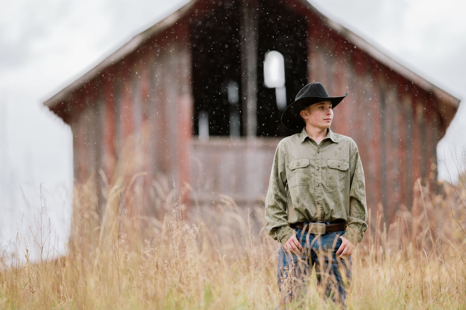 A person in a black hat and button down shirt stands in front of a weathered wooden barn surrounded by tall grass.