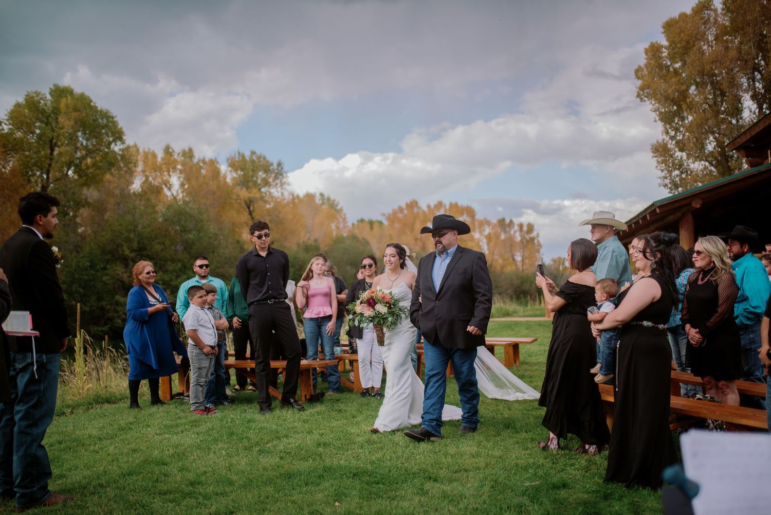 Rustic outdoor wedding ceremony with guests gathered on lawn near wooden benches.