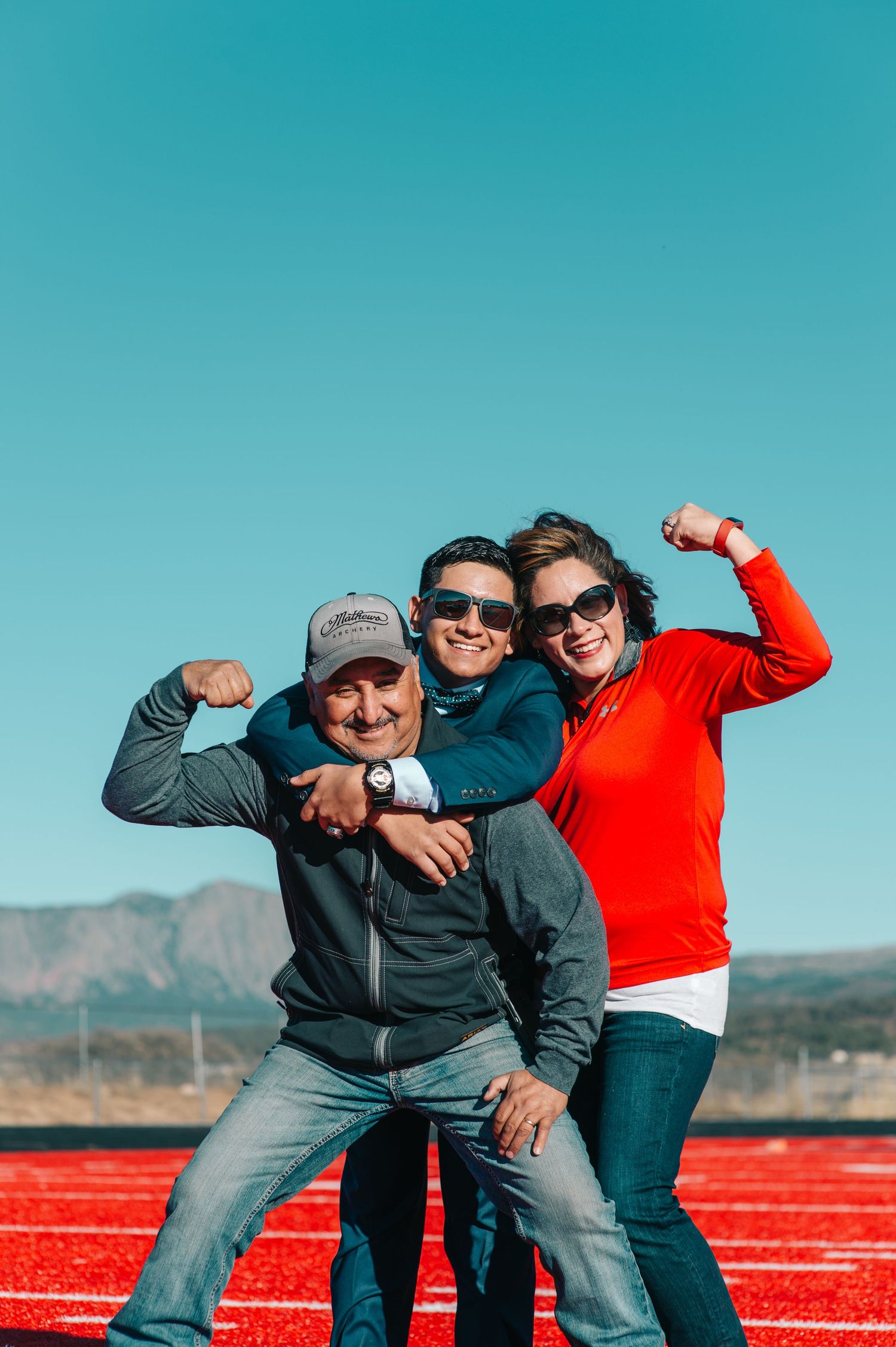 Three people flexing muscles on a red track with mountains and blue sky backdrop.