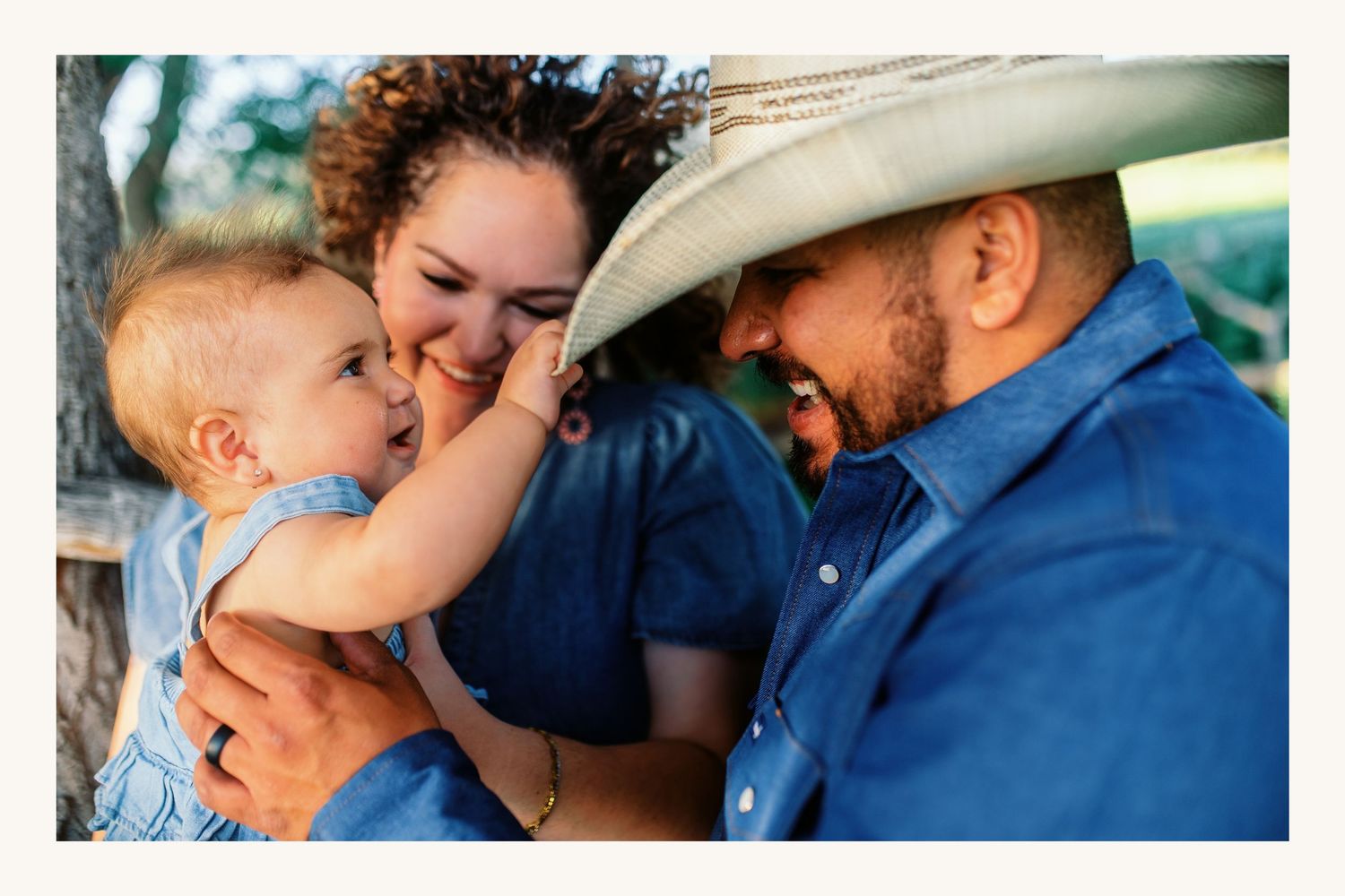 A young family shares a tender moment as a baby reaches toward dad in a cowboy hat outdoors.