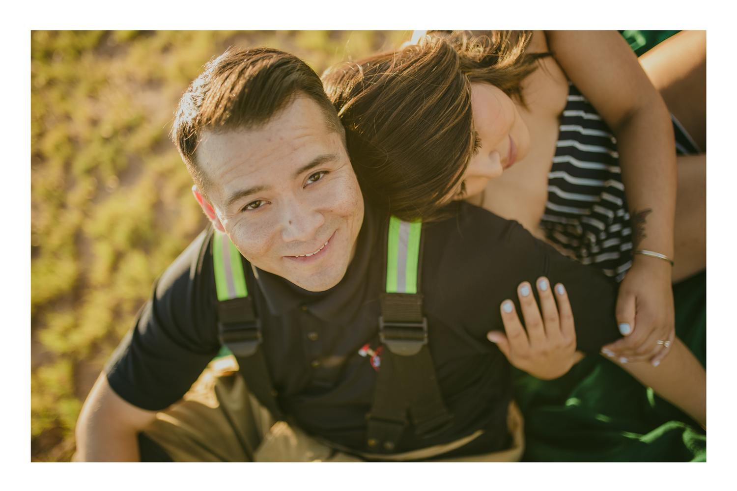 Young man in black overalls with green straps smiles up at camera outdoors in warm golden light.