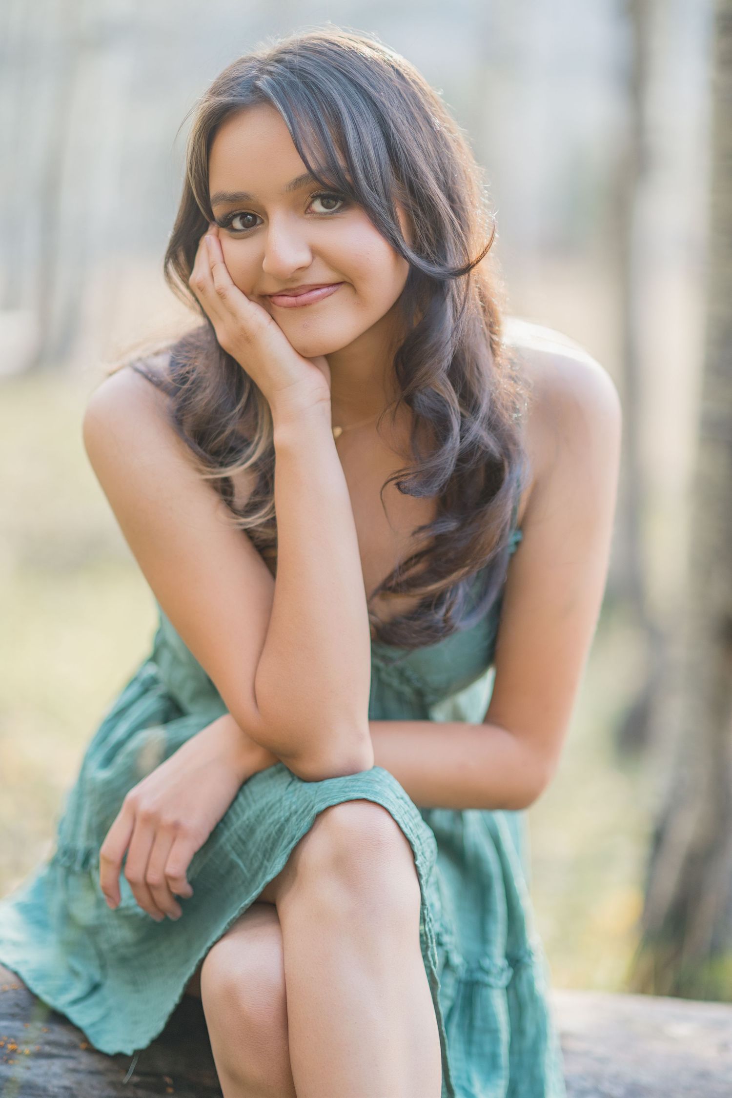 A person in a teal dress sits thoughtfully outdoors in a natural setting with soft lighting and blurred background.