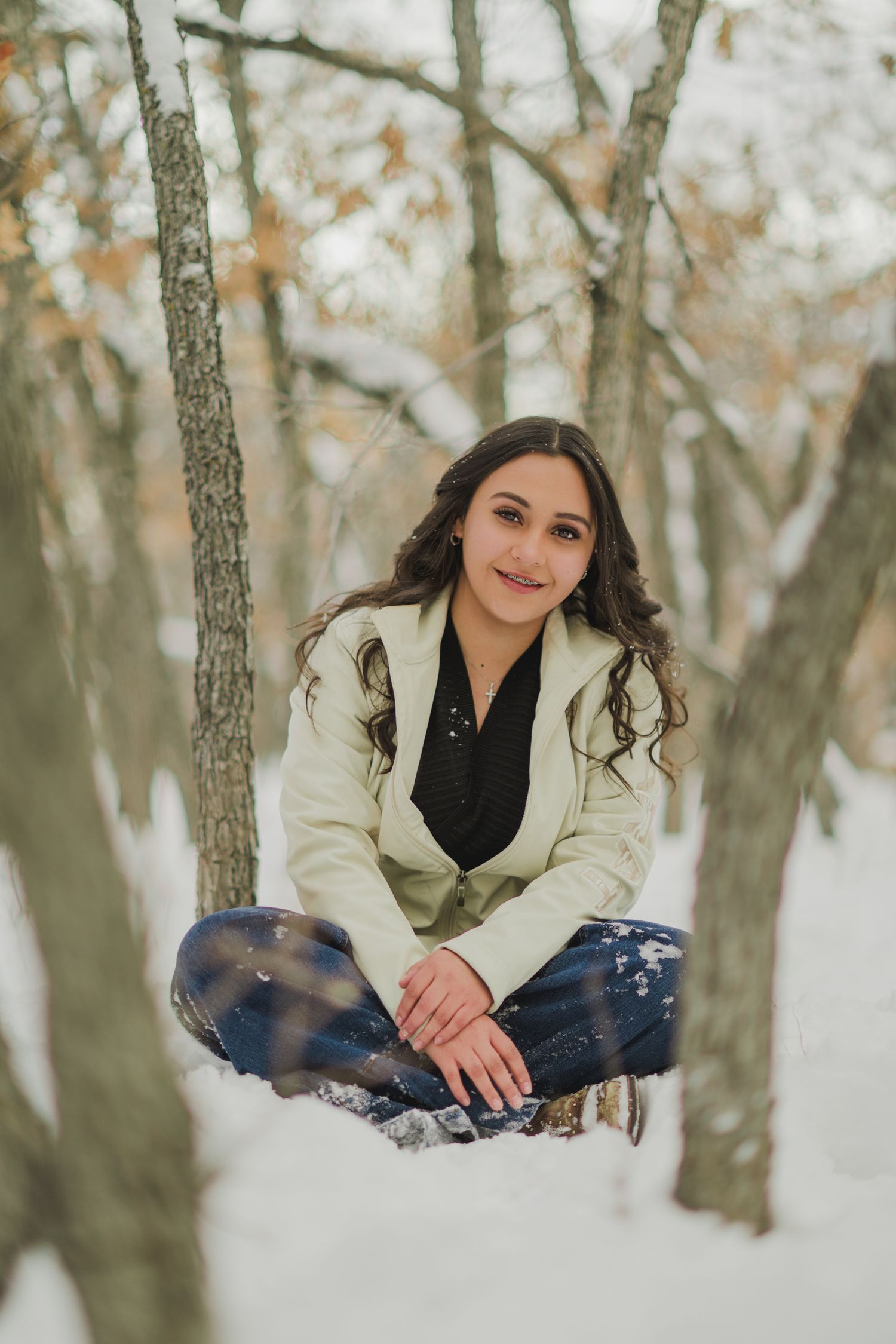 A person wearing a cream coat and black scarf sits in snowy winter woods during an outdoor portrait session.