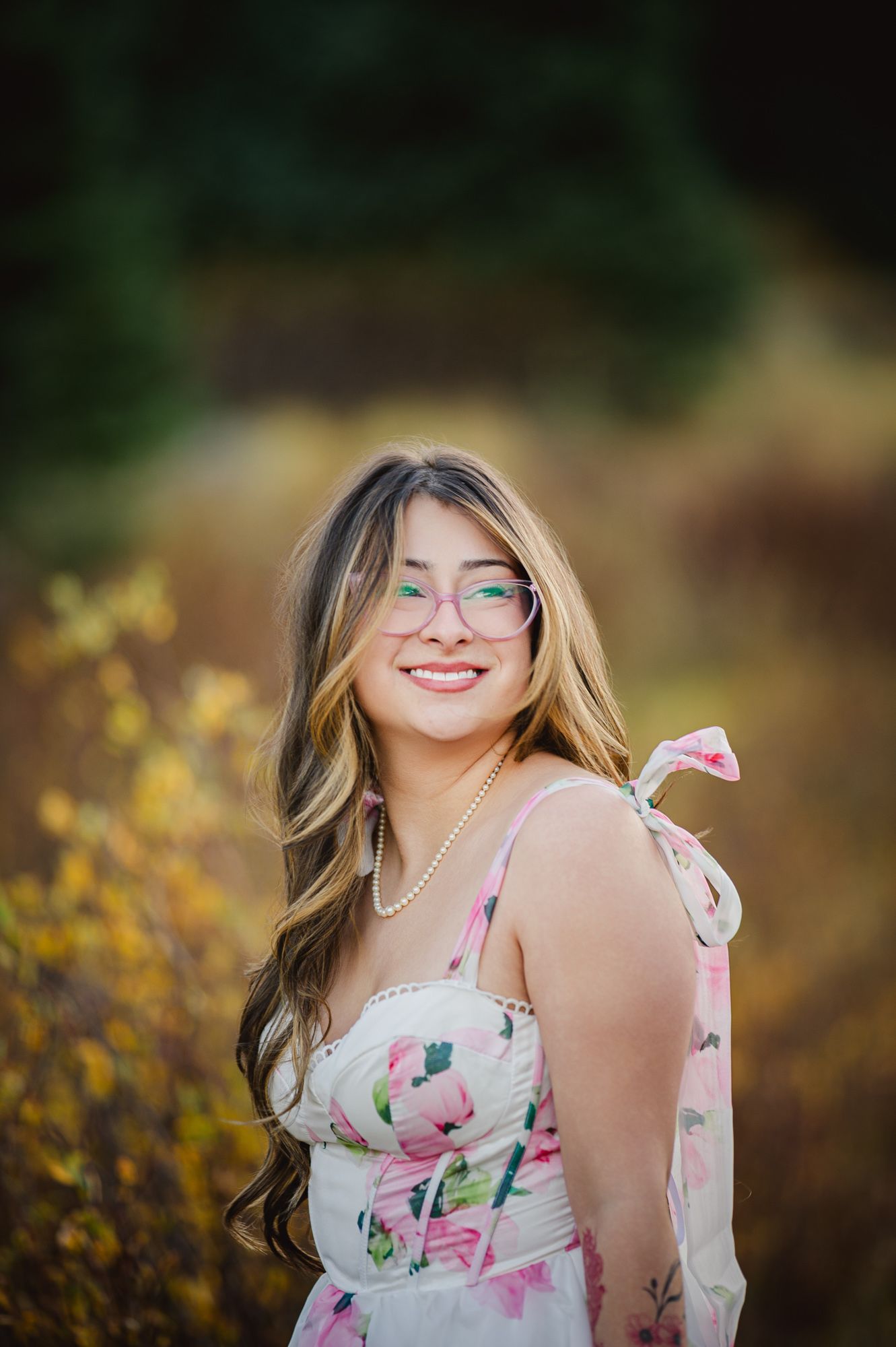 Bride-to-be in a floral dress and pearl necklace smiling among autumn leaves, Chama, NM engagement photos.