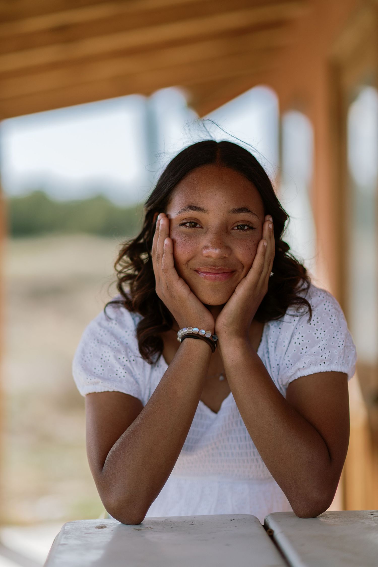 A person rests their head in their hands while sitting at a wooden table on a porch with natural lighting.