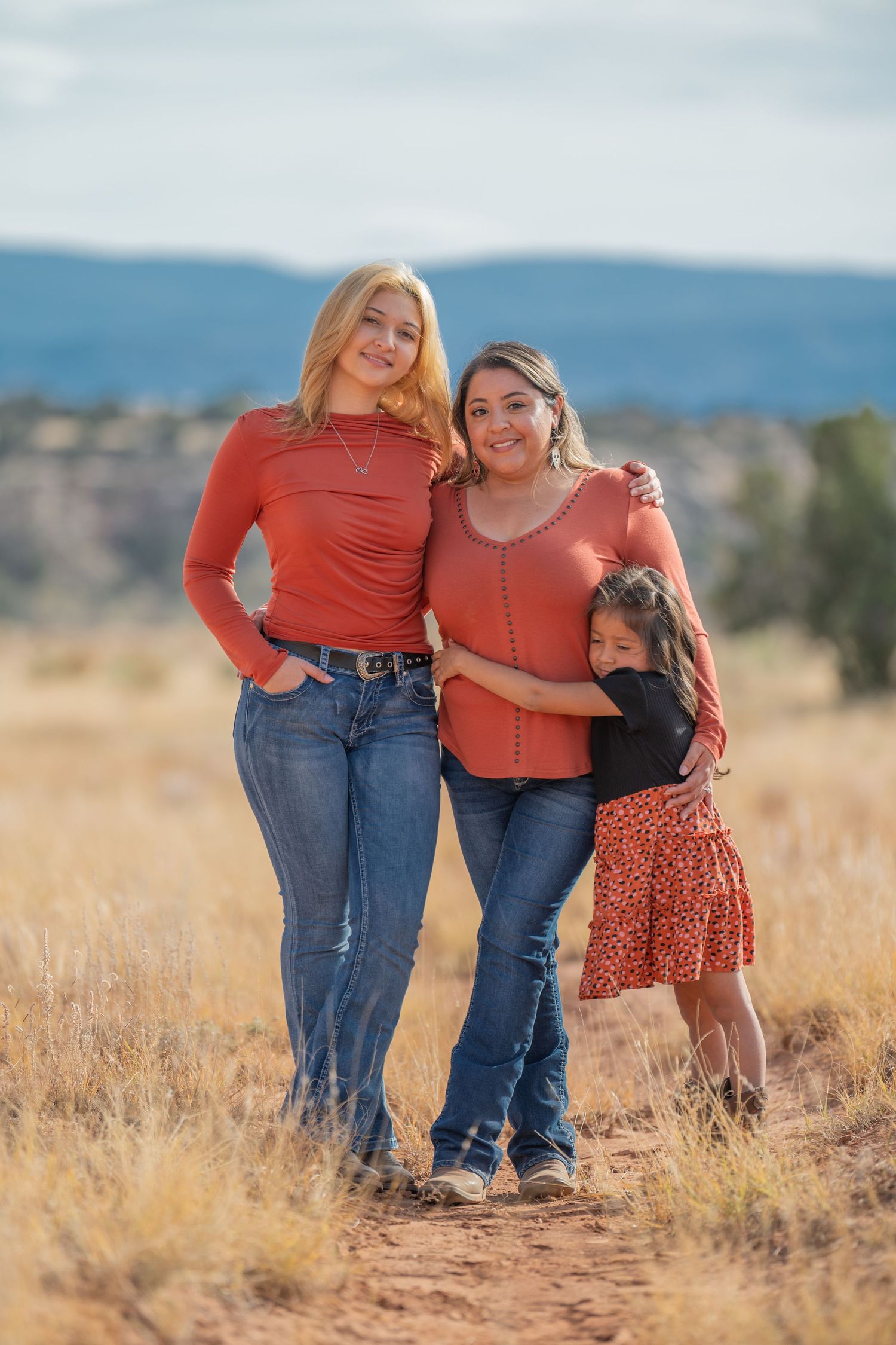 Three people wearing coordinating rust-colored tops pose together in a golden field with mountains in the background.