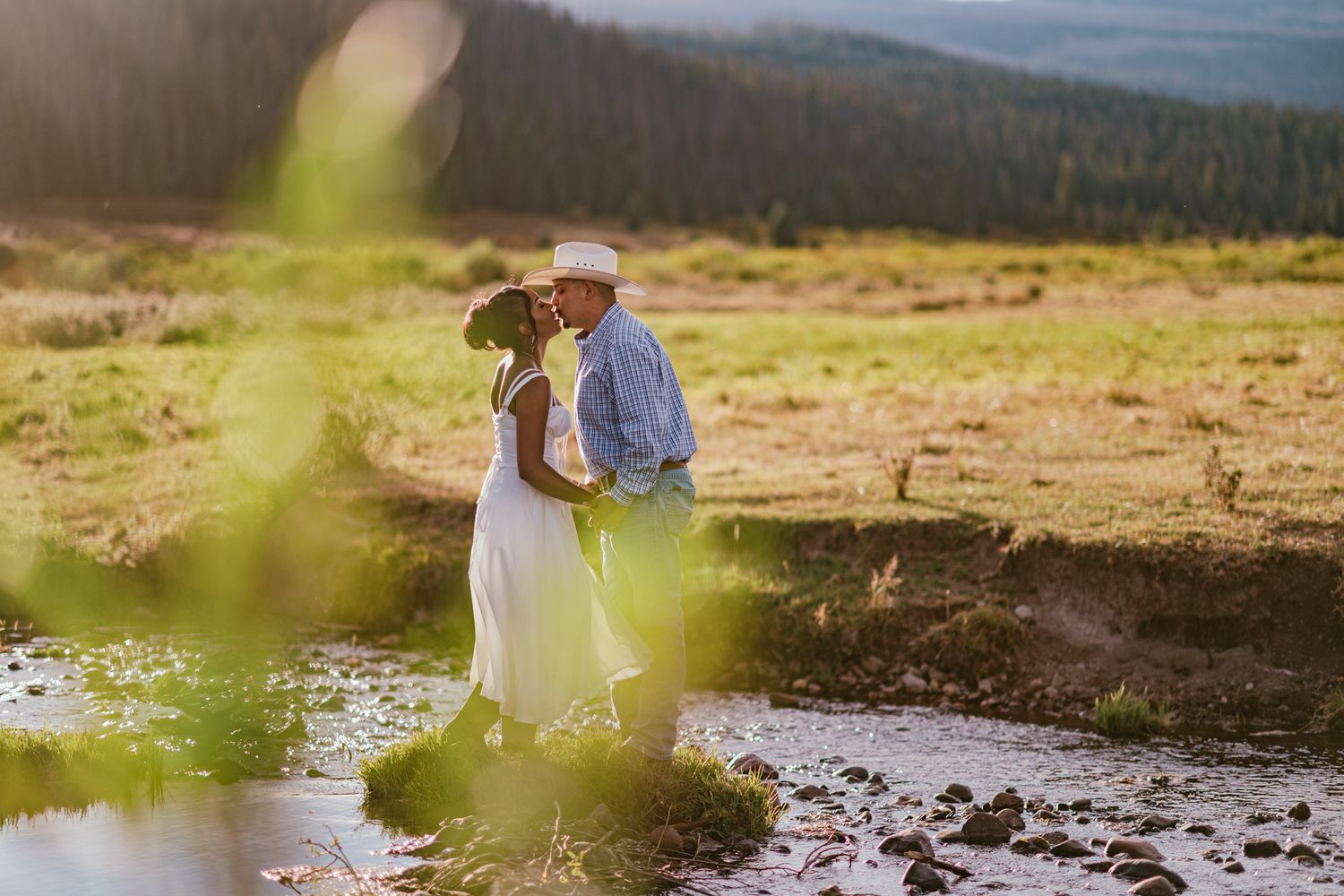 A couple shares a romantic kiss in a sunlit mountain meadow, bride in white dress and groom in hat near a stream.