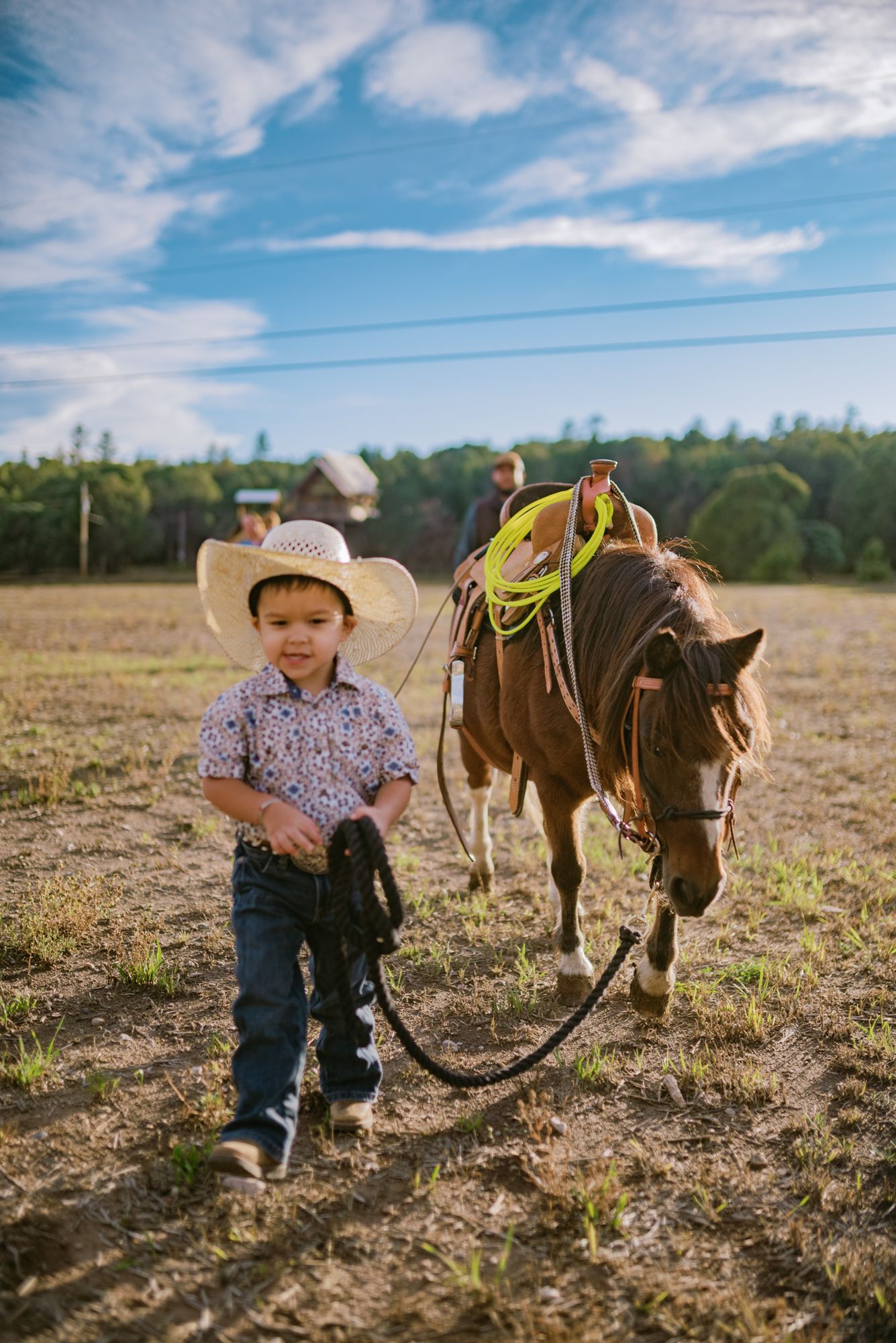 Young boy in cowboy hat and boots leading a brown horse across a sunny ranch field.