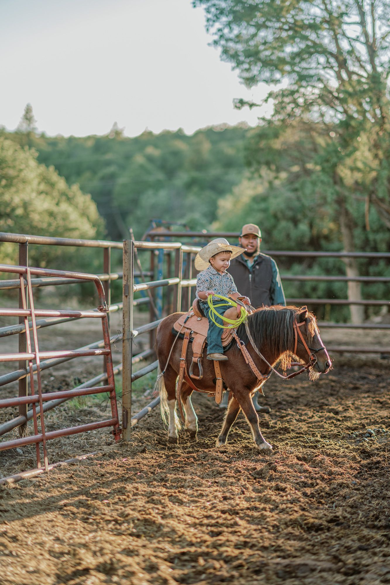 Young child in cowboy hat riding a brown pony in a ranch corral with an adult supervising nearby.
