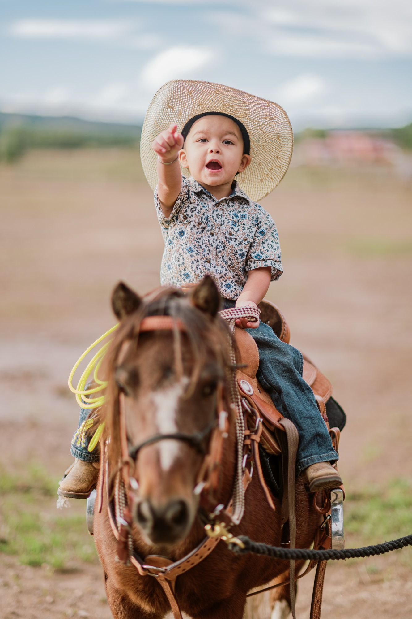 Young child in cowboy hat and boots riding a brown horse outdoors, pointing up with one hand.