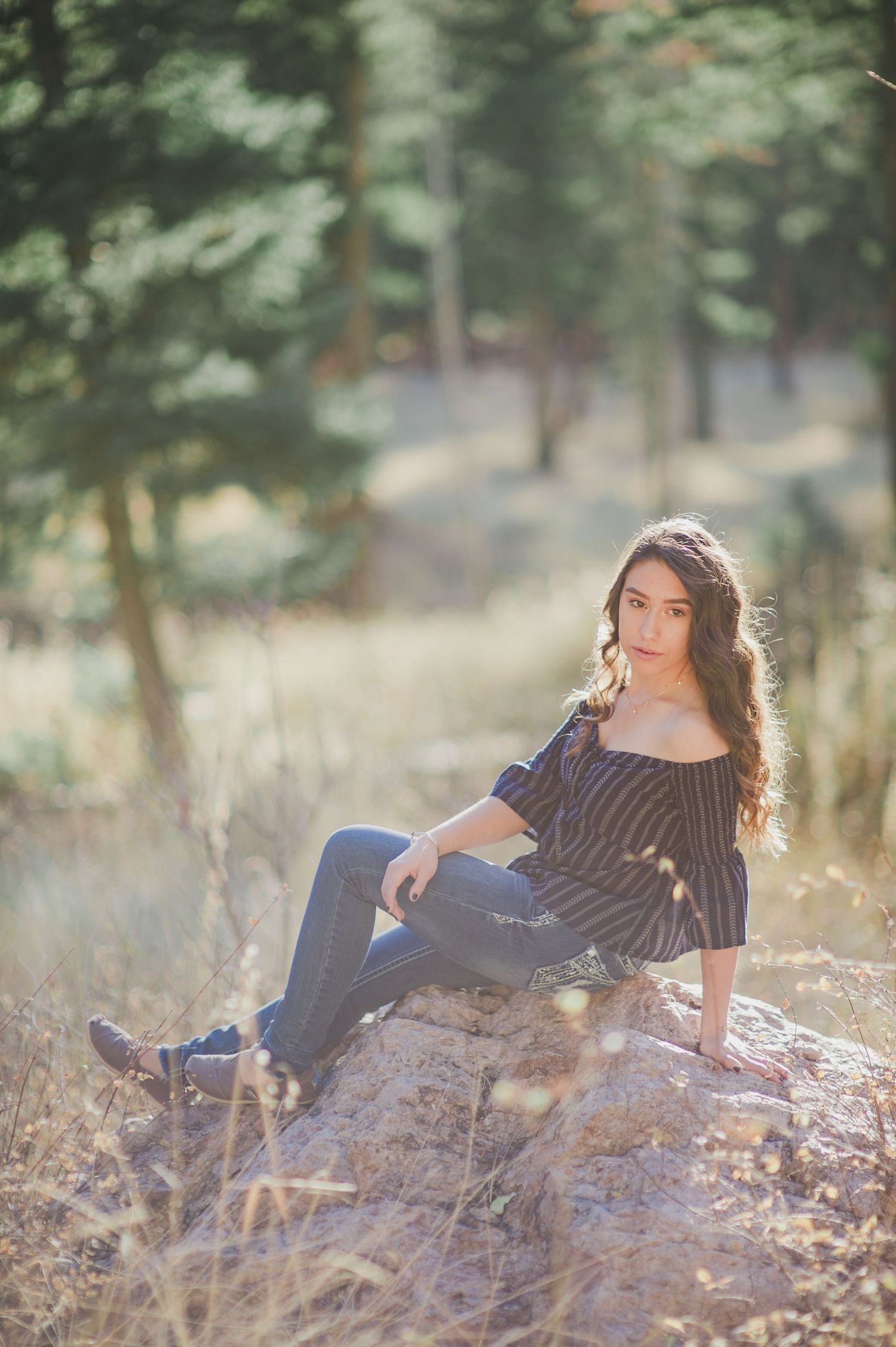 Young woman in striped off-shoulder top and jeans sitting on a rock in a sunlit forest meadow.