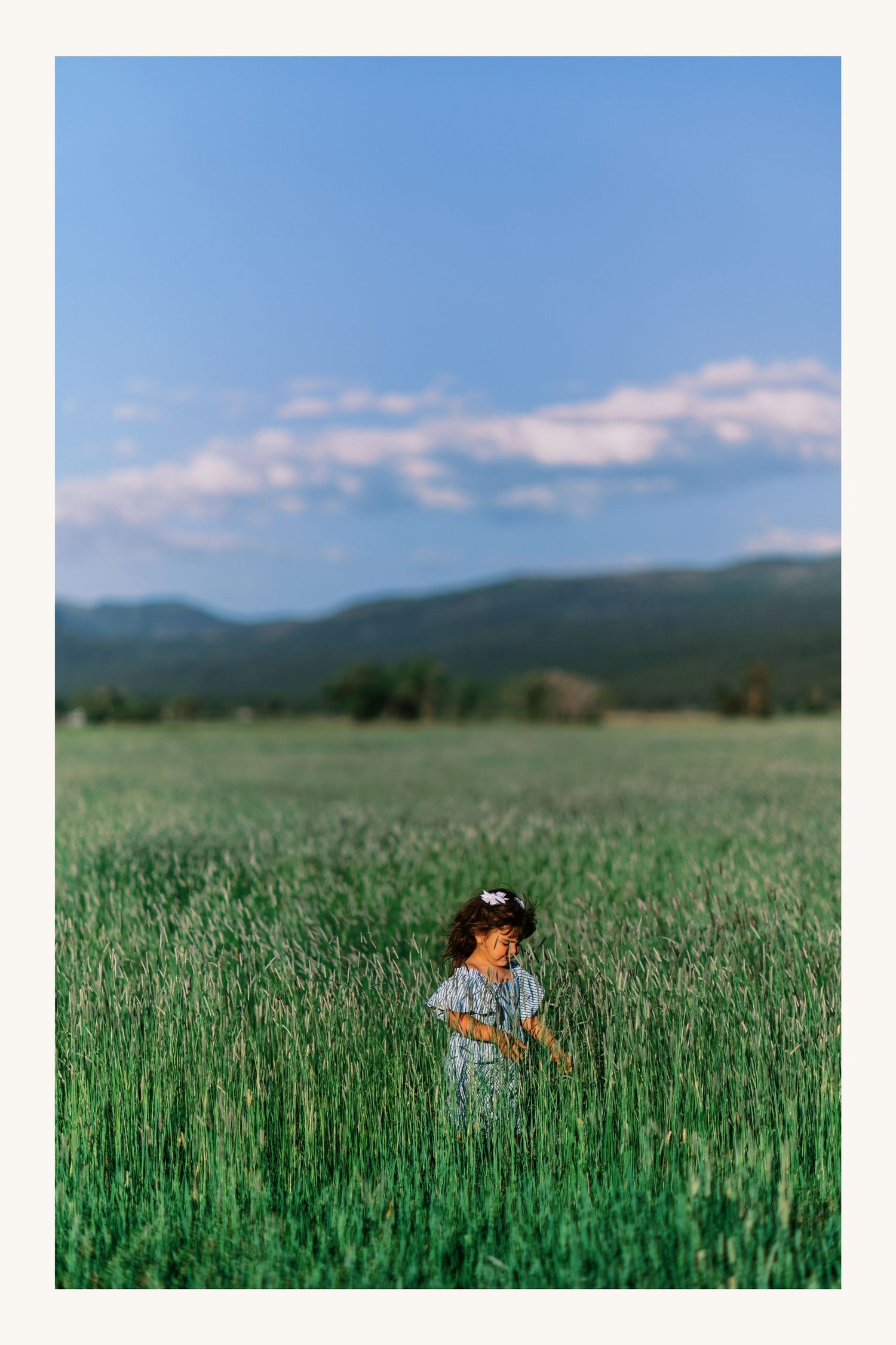 A young girl in a floral dress stands in a vast green field under a blue sky with mountains in the distance.