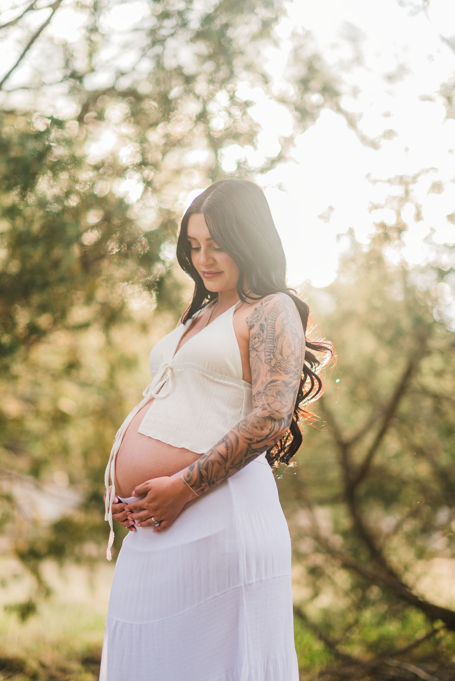 Pregnant woman in white dress with tattoed arm cradling baby bump outdoors in golden sunlight.