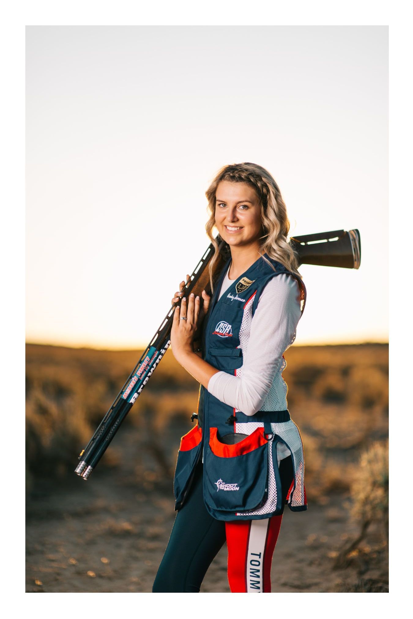 Female competitive shooter in USA vest holding shotgun outdoors at sunset in desert landscape.