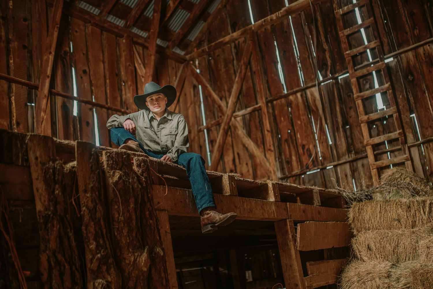 A rustic barn interior scene with someone sitting on wooden stairs next to hay bales in atmospheric lighting.