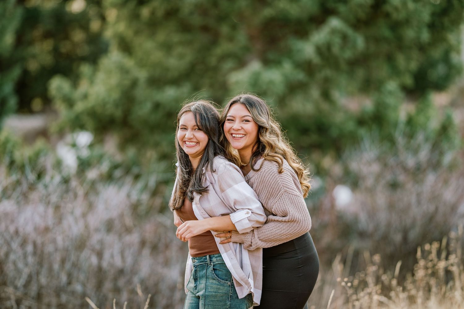 Two friends stand together laughing and embracing in an outdoor setting with wildflowers and natural greenery.