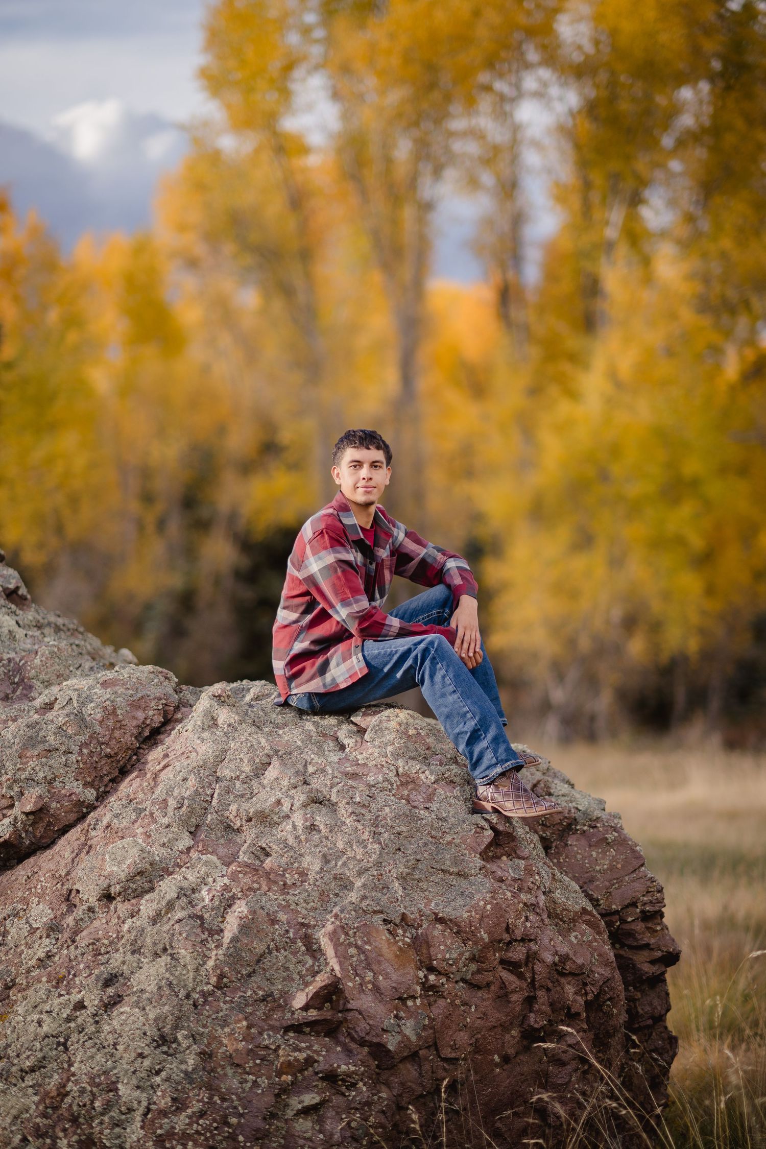 Person in plaid shirt and jeans relaxing on large rock formation with golden autumn trees in background.