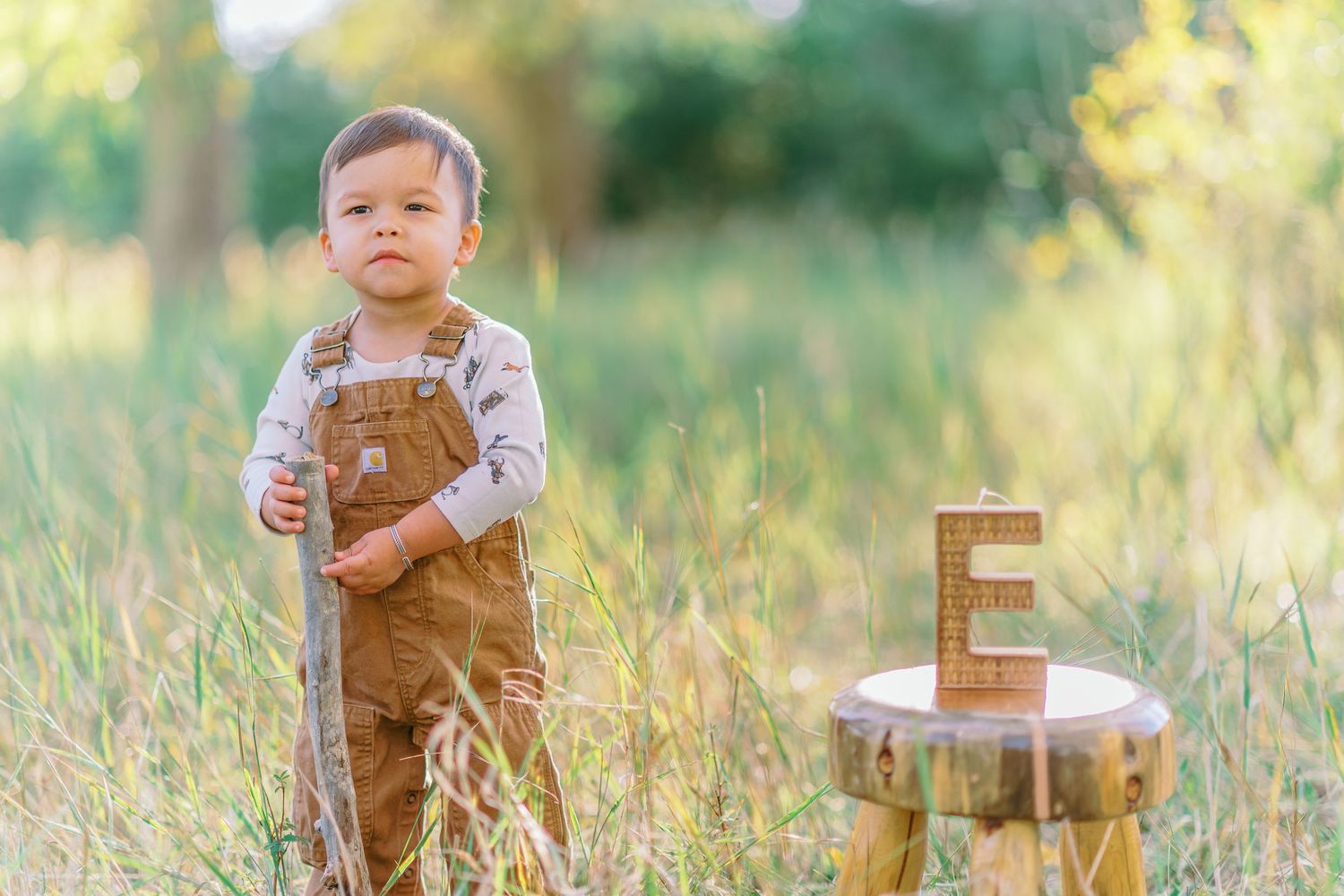 A toddler in brown corduroy overalls plays in a scenic field during golden hour while holding a wooden letter block.