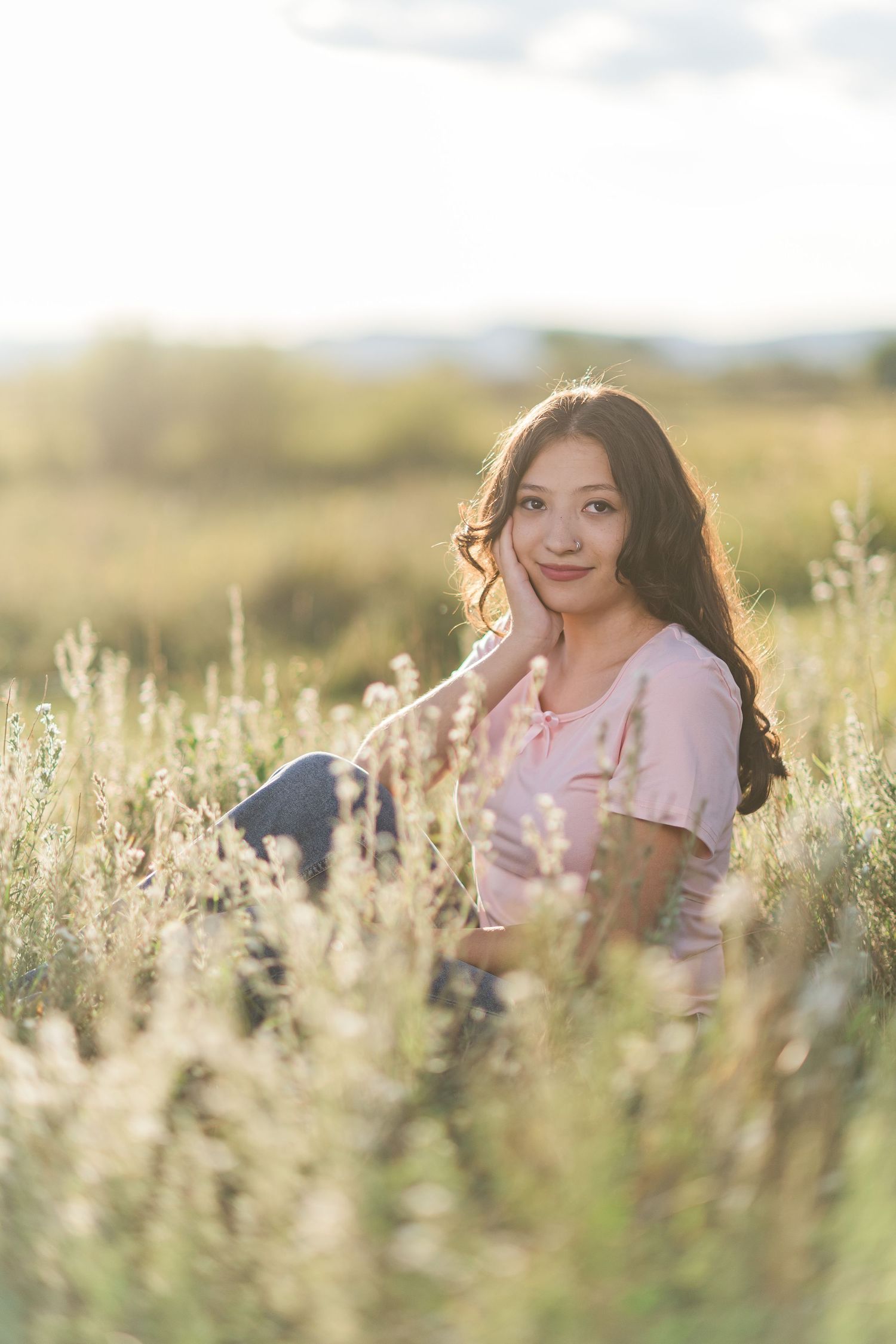 A person in a pink top sits among tall wildflowers in a sunny field during golden hour.