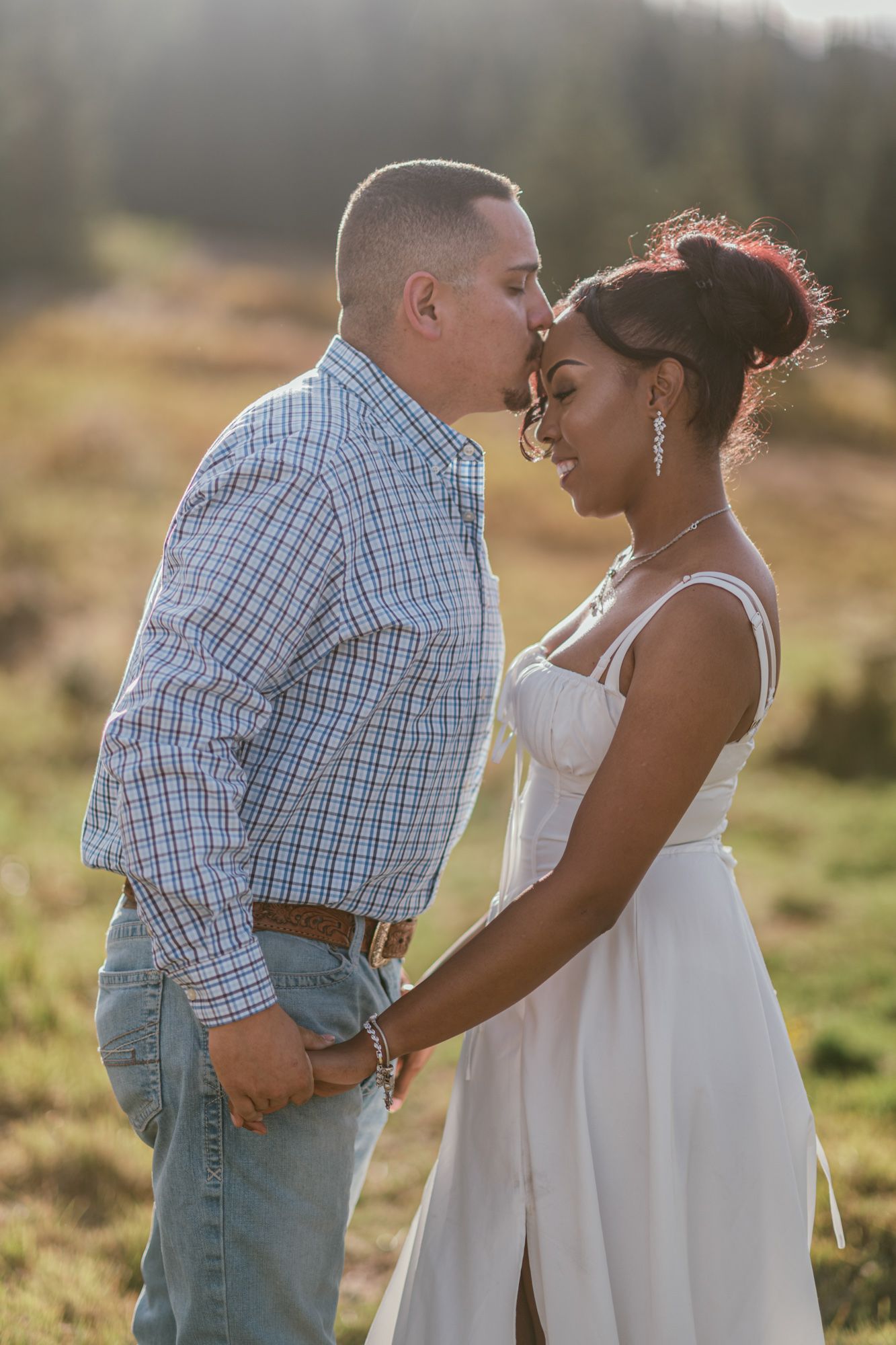 Couple sharing a tender forehead kiss in a sunlit meadow, man in plaid shirt and woman in white dress holding hands.