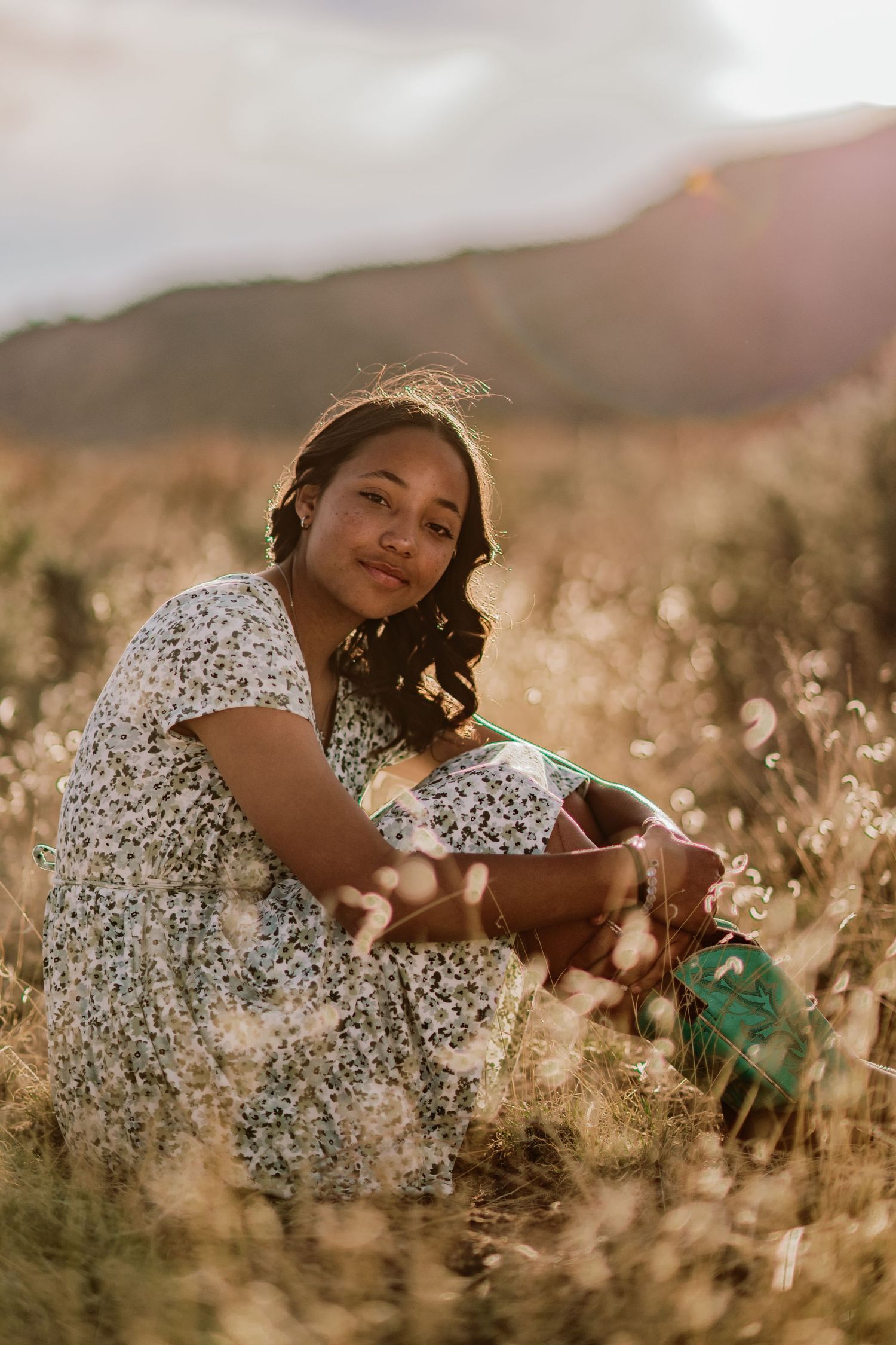 Someone in a floral dress laying in a golden sunlit field of tall grass and wildflowers at sunset.