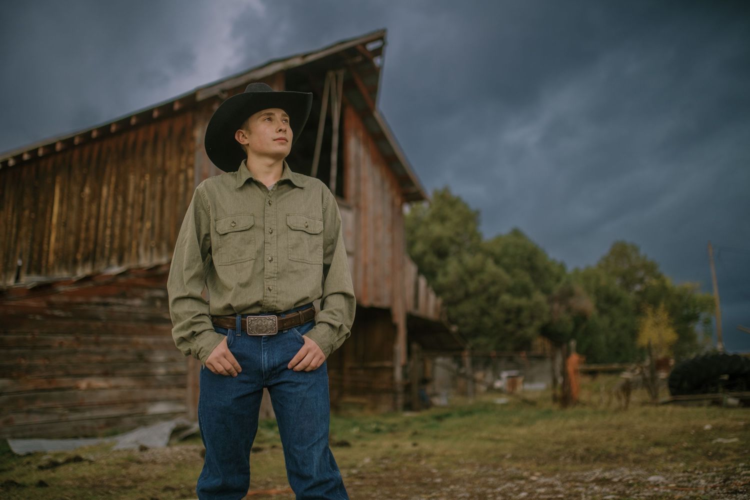 Vintage barn setting at sunset with dramatic storm clouds and figure in western attire creates moody countryside ambiance.