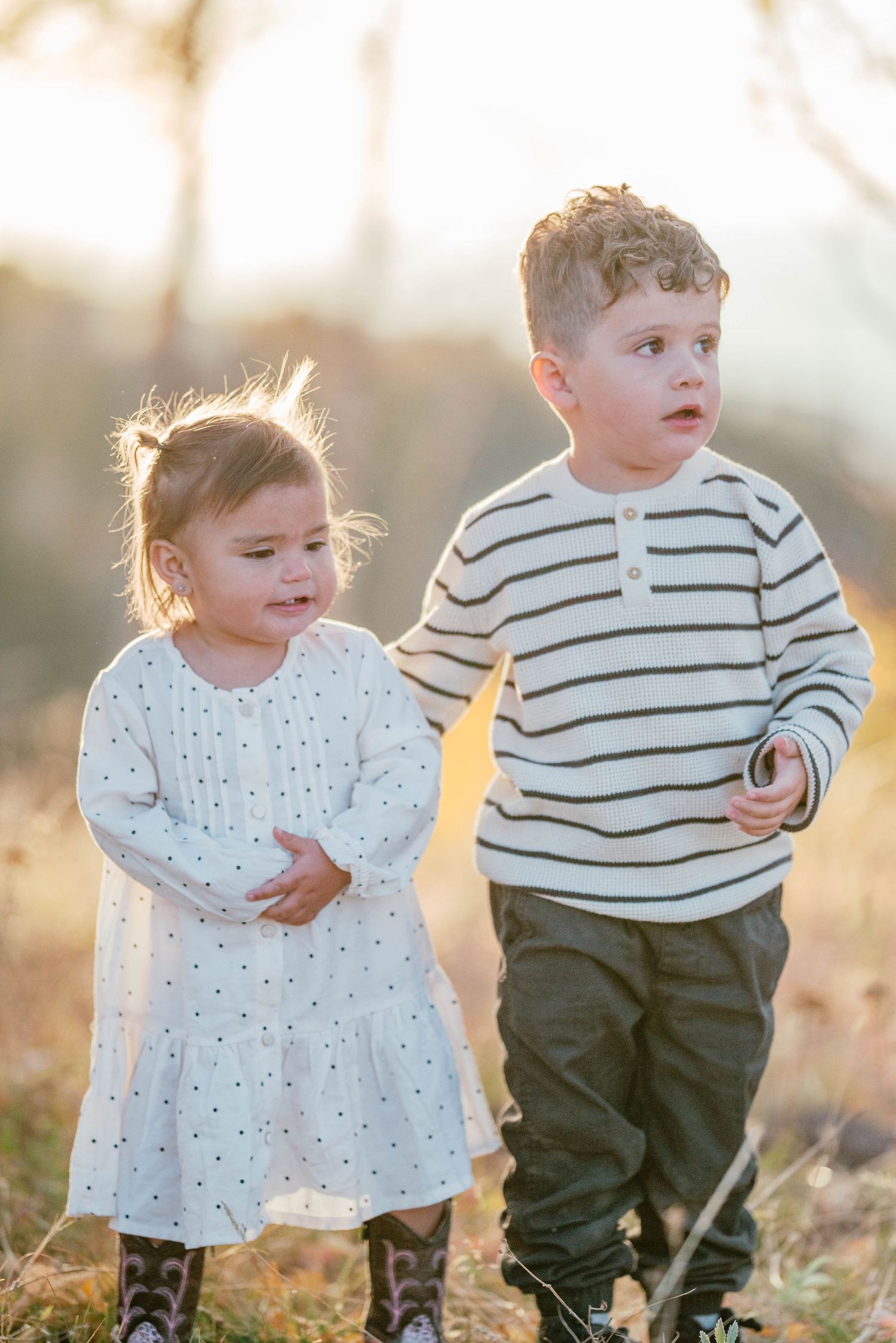 Two young children in backlit outdoor setting wearing casual clothing stand together at sunset.