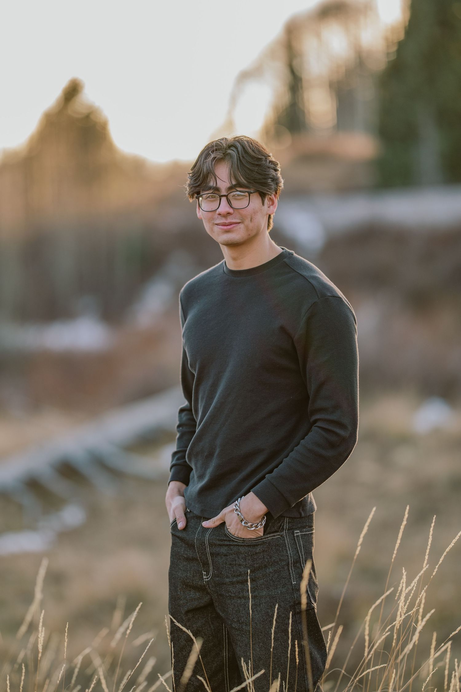 Person in black sweater and glasses poses in golden field during sunset with blurred background.