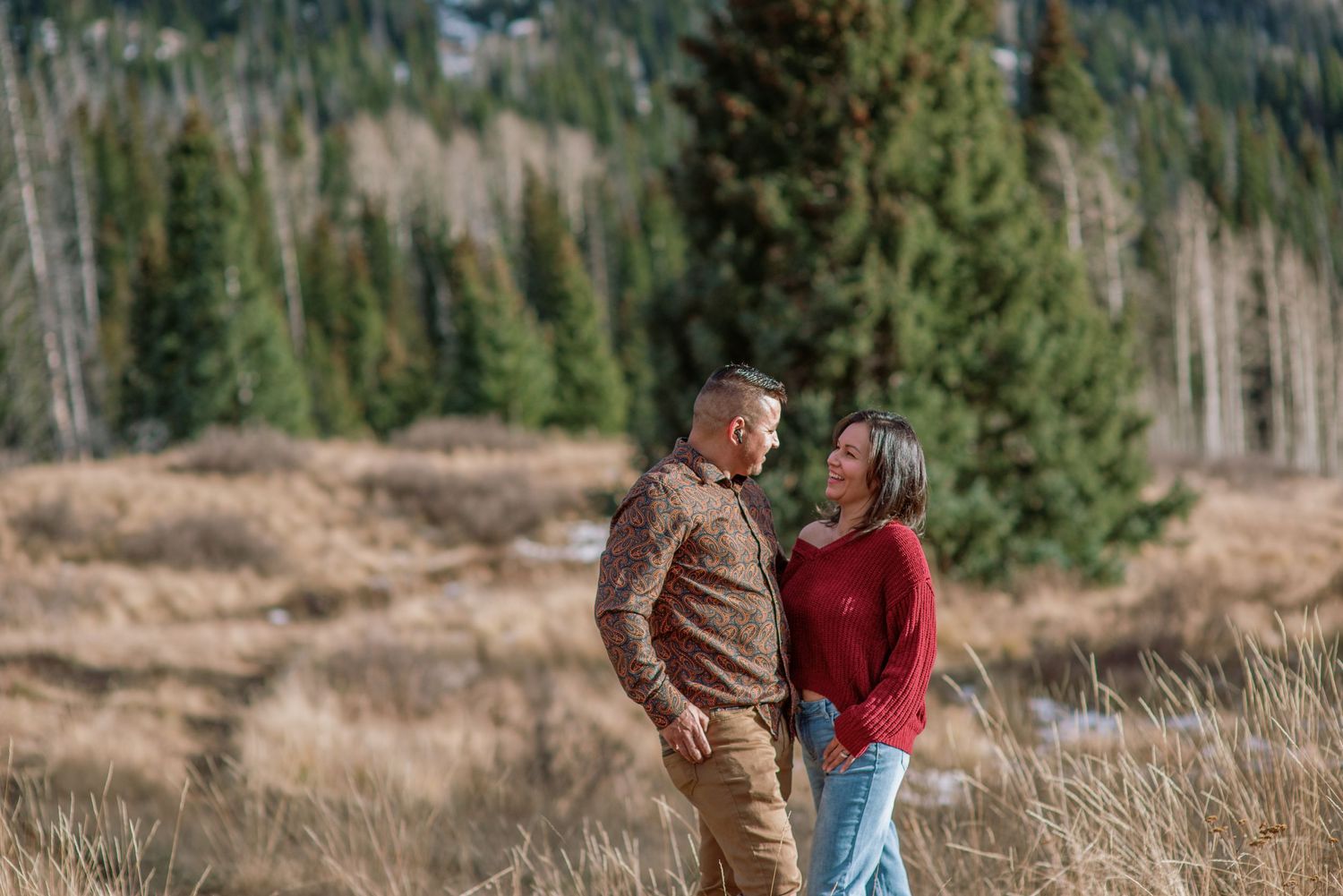 Couple in brown and red jackets sharing a romantic moment in a scenic mountainous forest landscape.